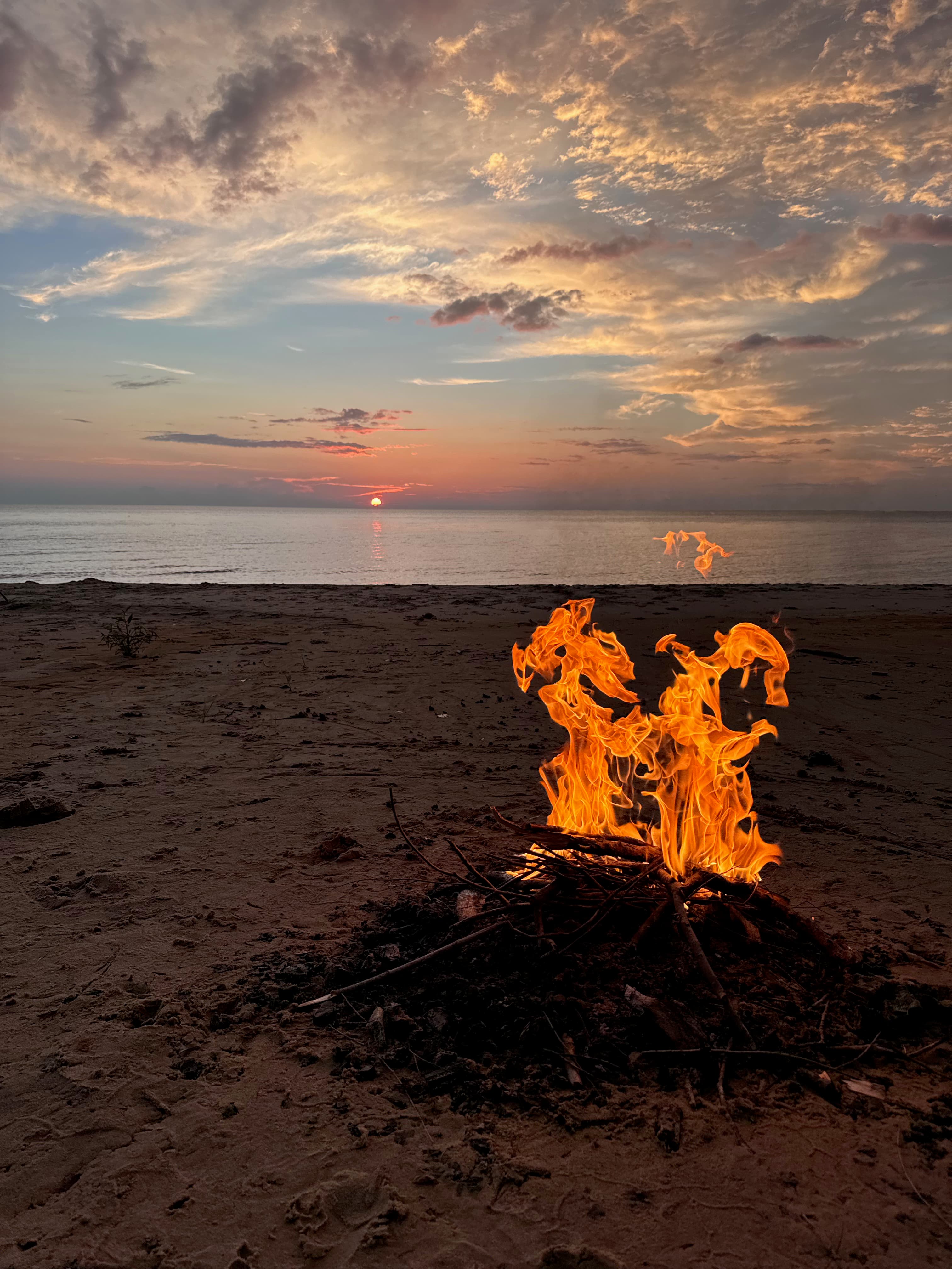 A picture of a campfire on the beach at sunset with a partly cloudy sky.