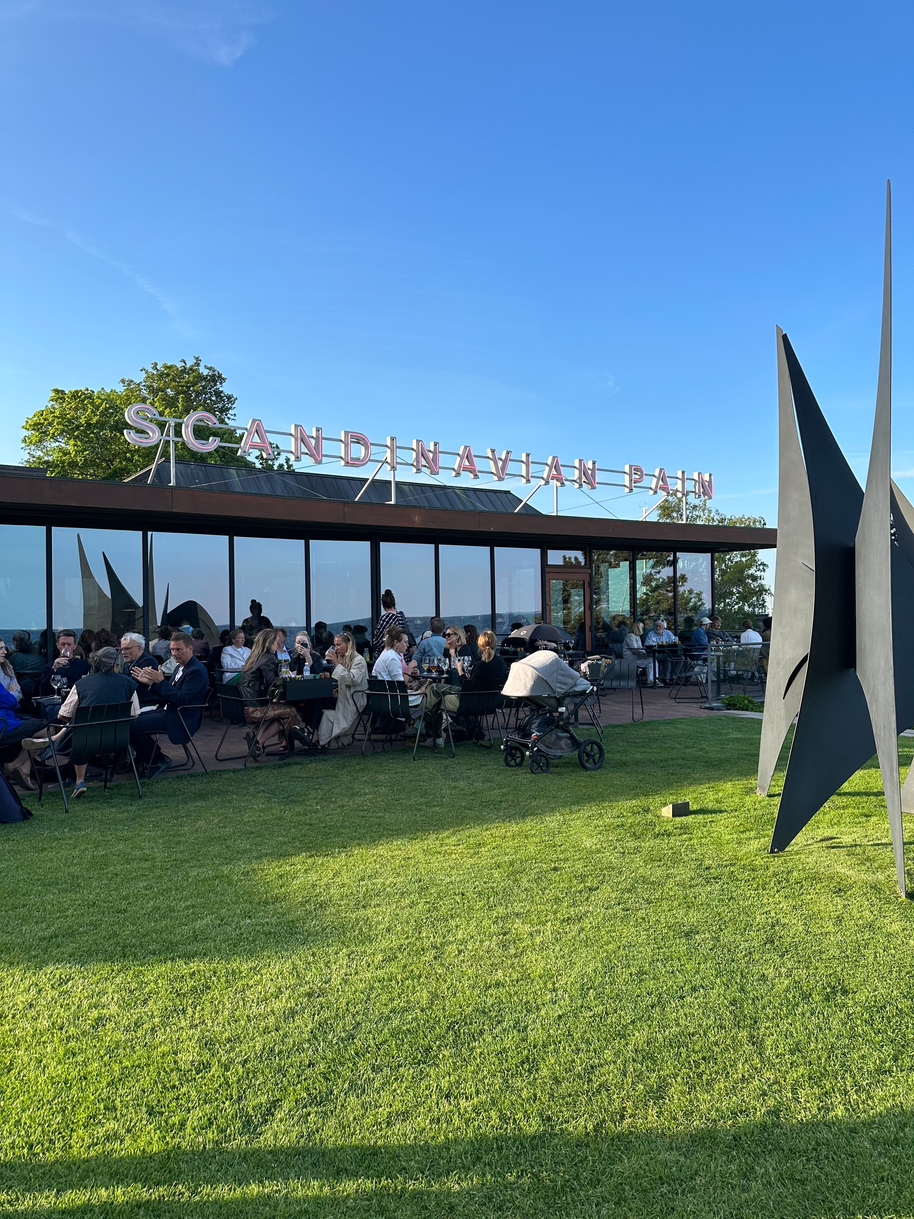 A grassy outdoor area with people sitting at tables by a cafe on a sunny day