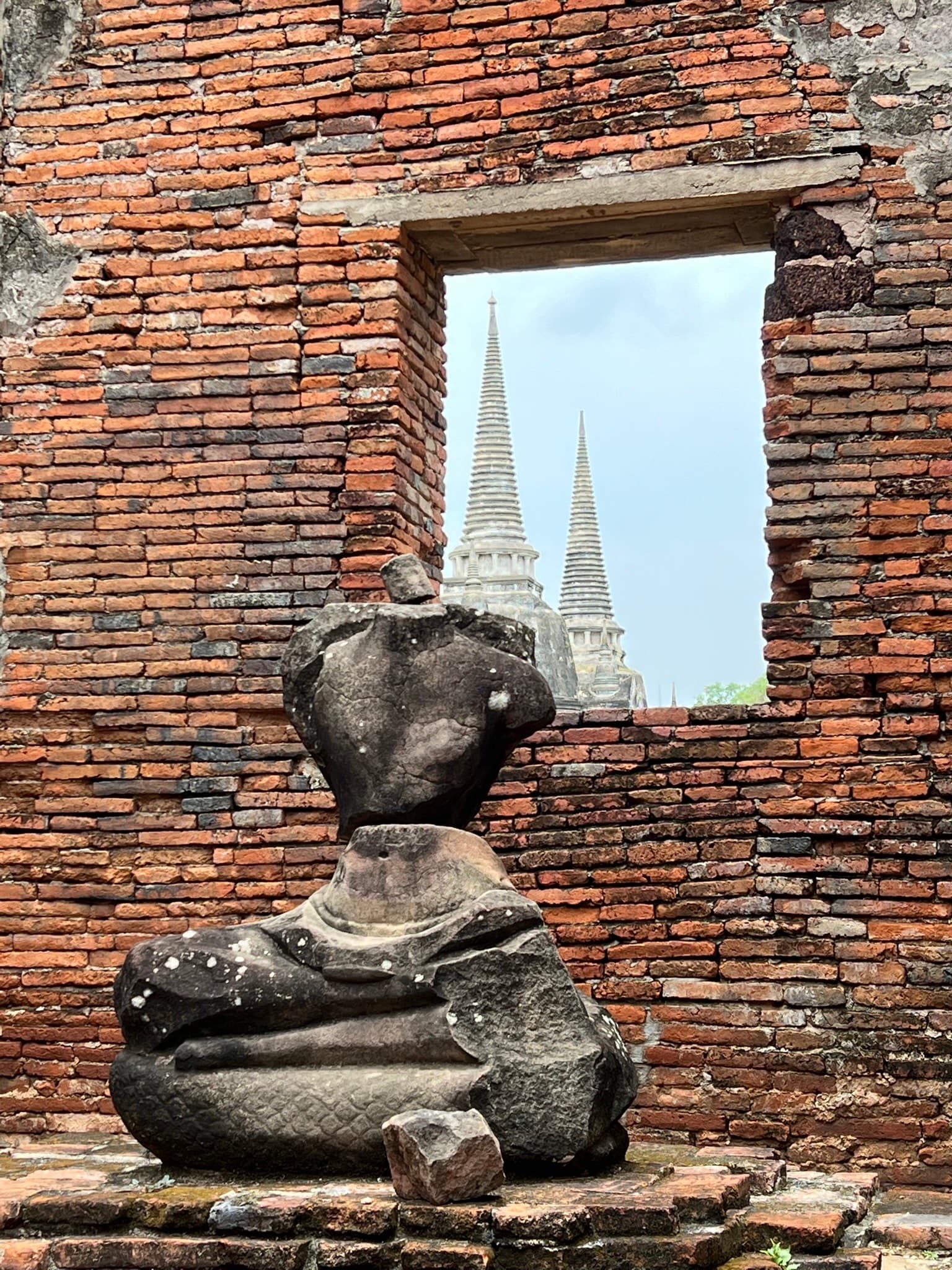 View of a headless seated statue by a brick wall with a temple spire visible though an opening