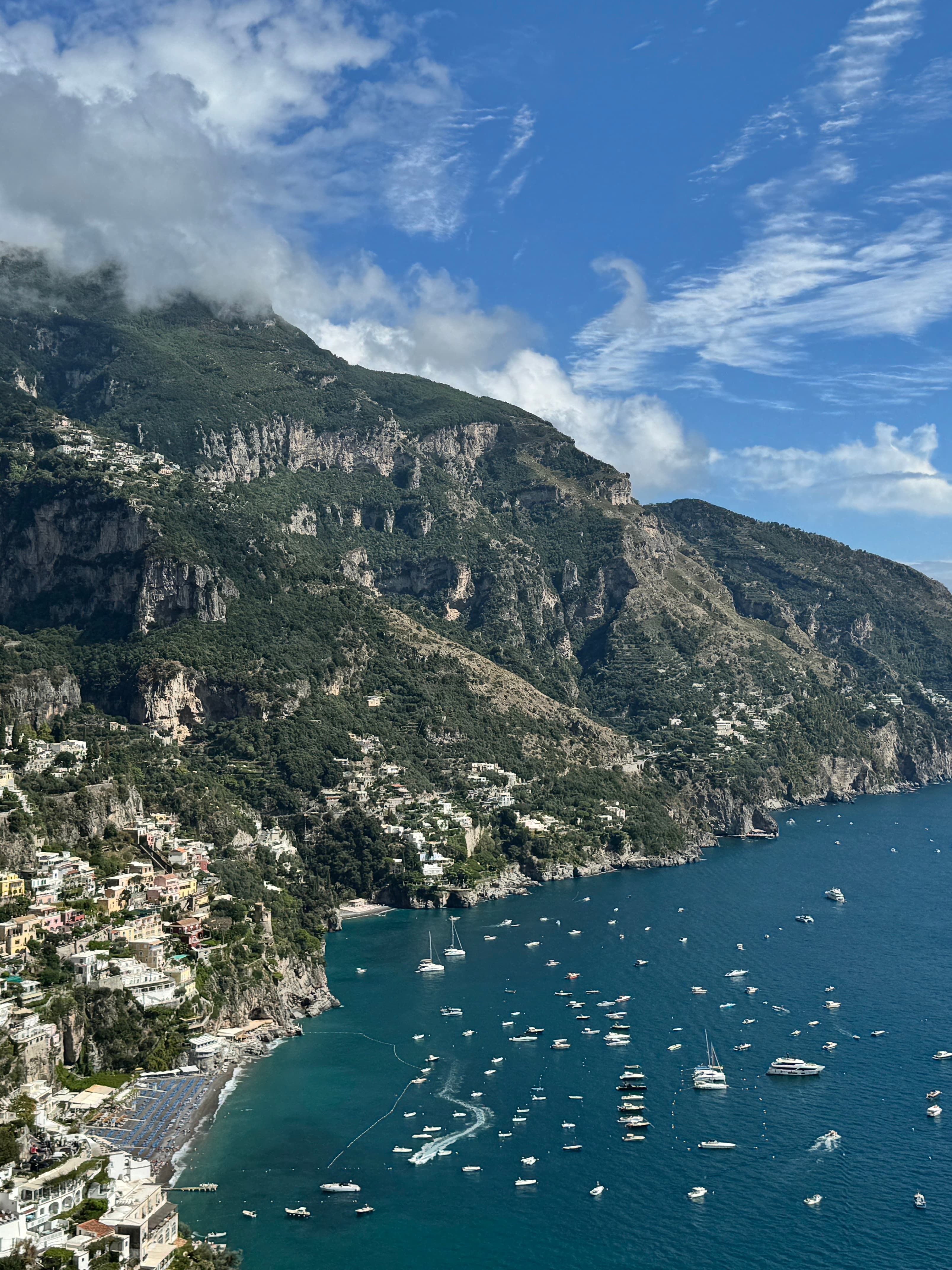 Aerial view of a coastline with green mountains and dozens of small boats anchored offshore on a sunny day