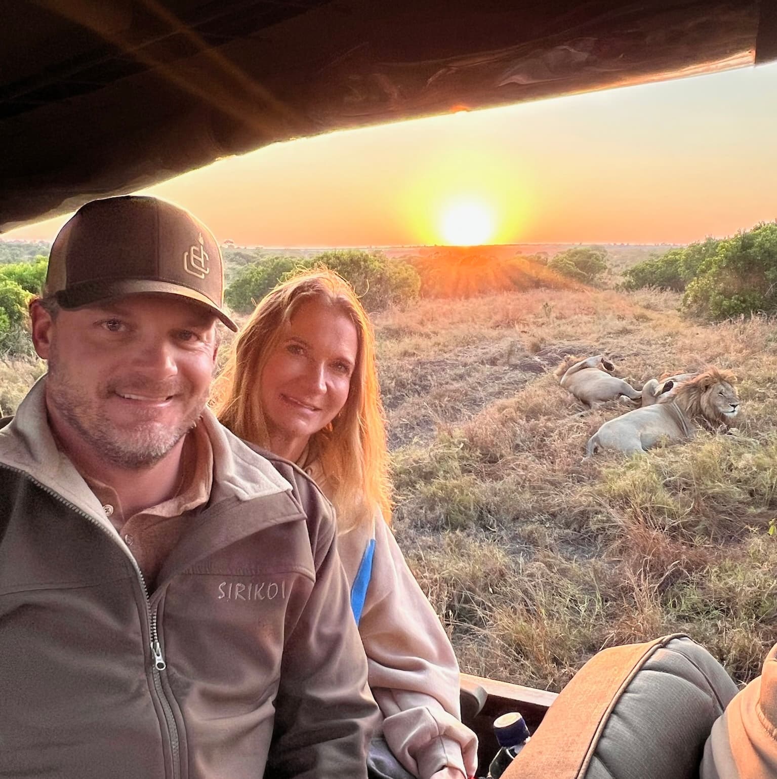 Advisor and partner smiling side by side in a safari vehicle at sunset with lions resting in the grass nearby