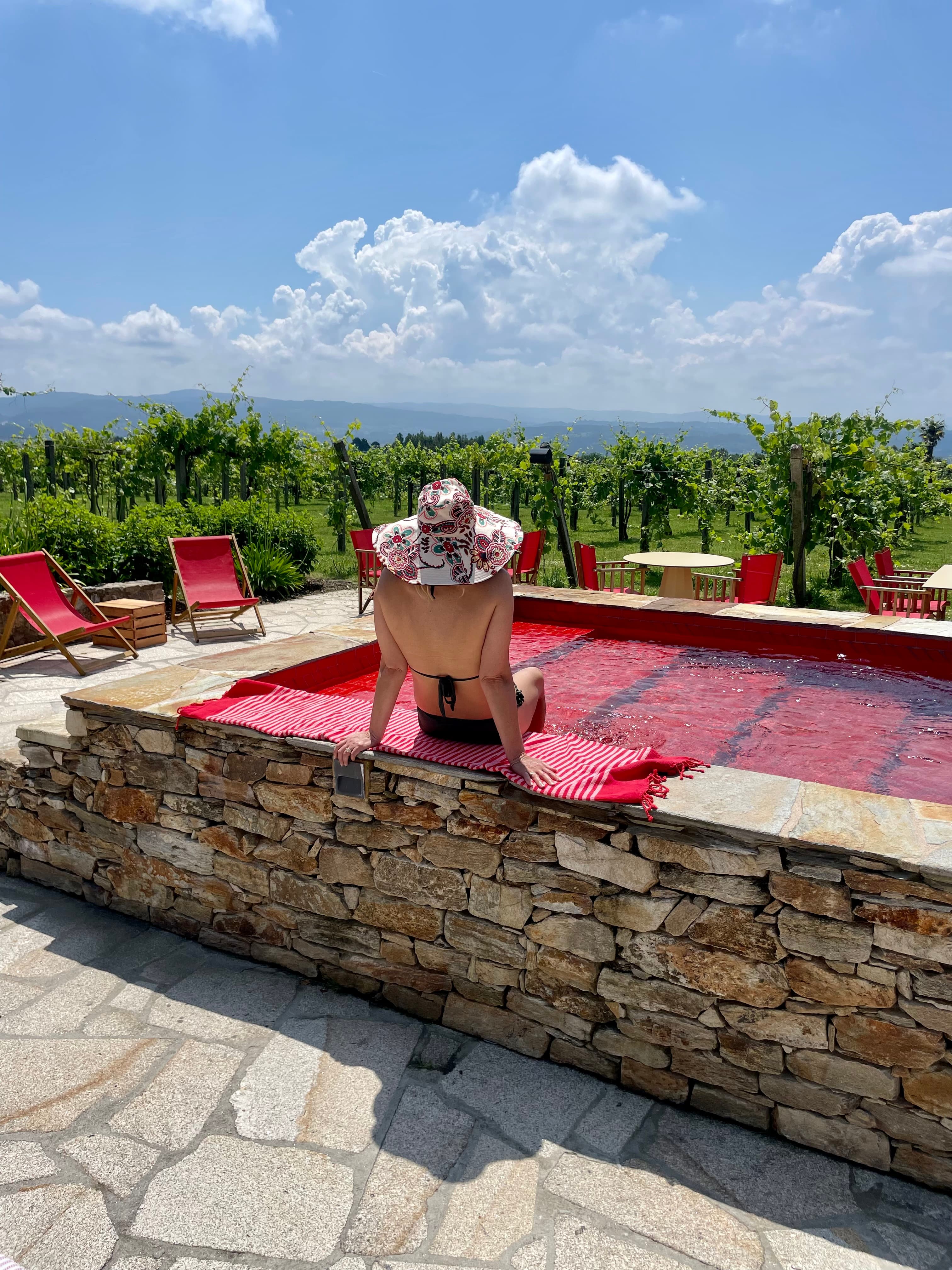 An advisor wearing a swimsuit and posing on the edge of stone wall with her feet dangling in a swimming pool that appears to have red tile. There are red lawn chairs and trees in the surrounding area.
