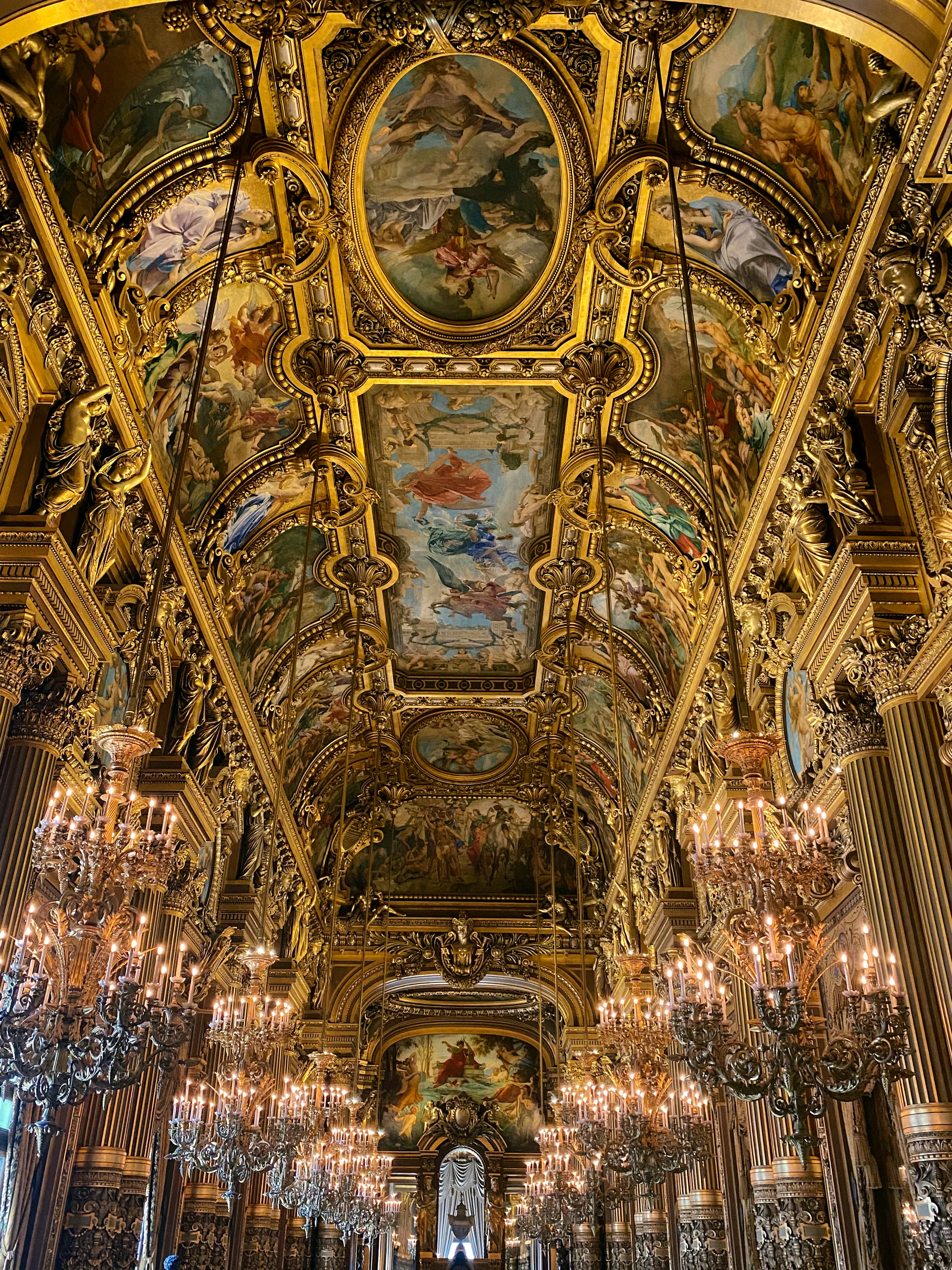 Interior view of ornately painted ceilings in a grand golden hall with rows of chandeliers hanging to either side