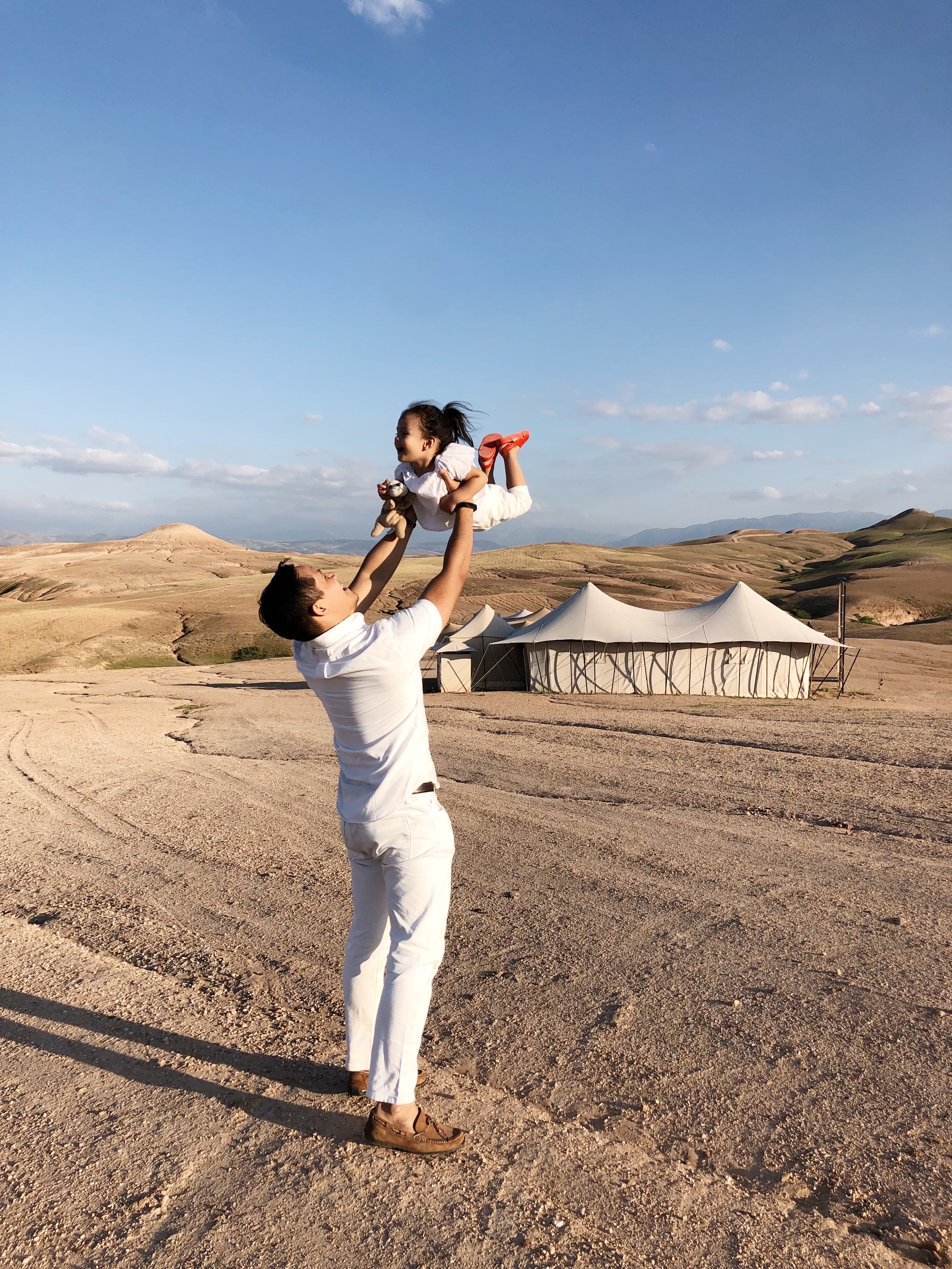 A man holding a young girl up in the air both dressed in all white in the desert under clear skies