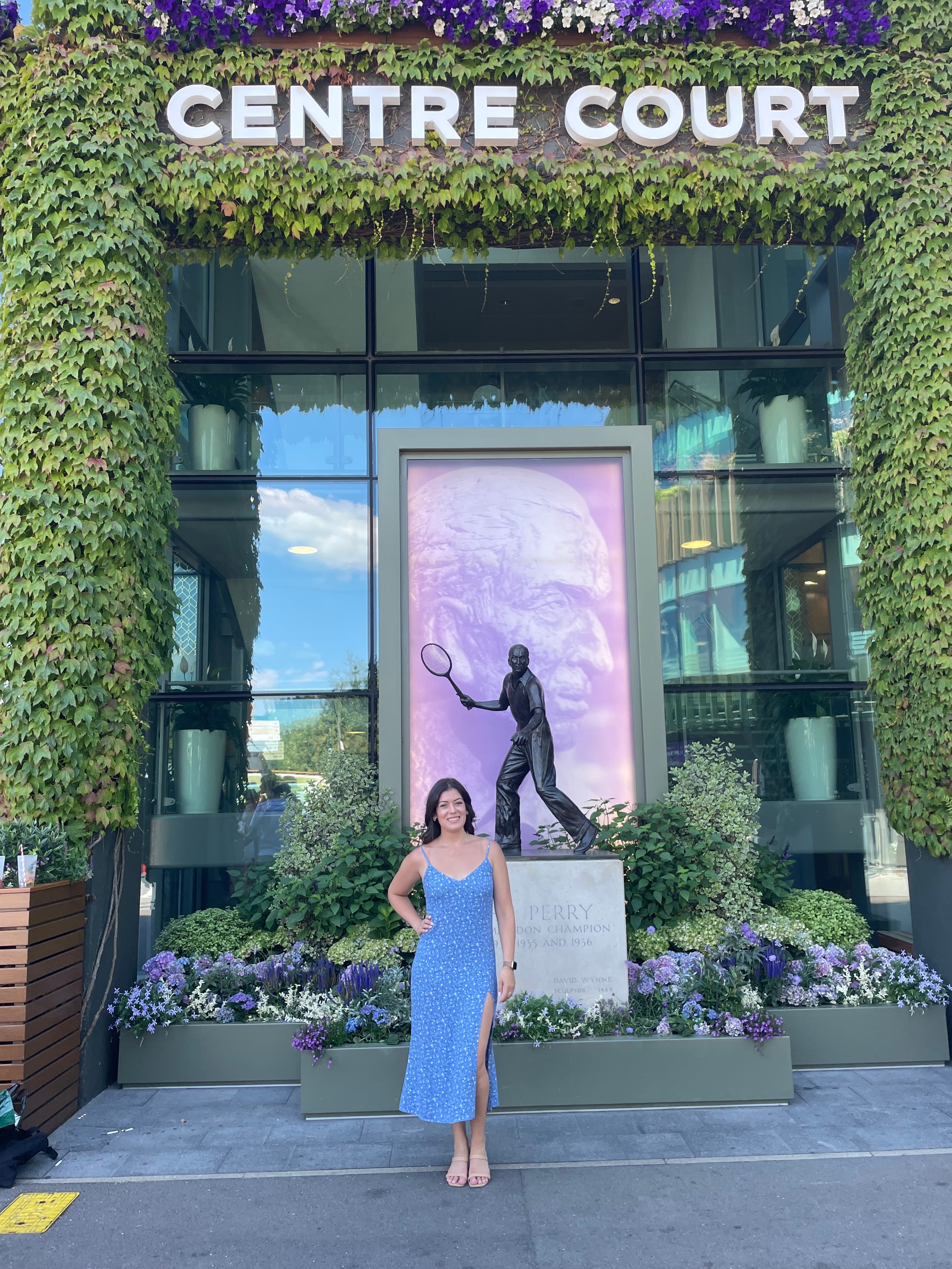 Jessica posing in a blue dress outdoors in front of a building with greenery around it.