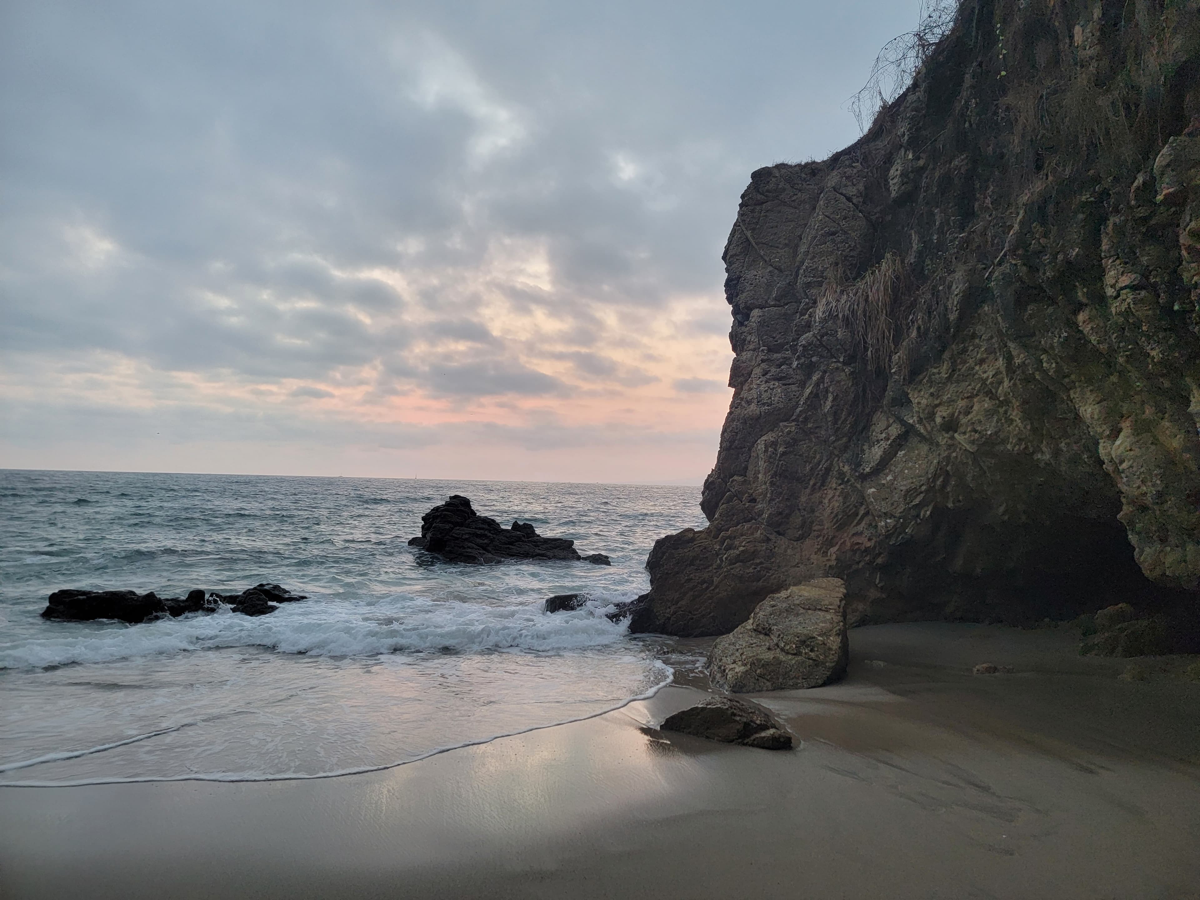 A view of a rocky cliff and beach with the sun lightly setting in the background.
