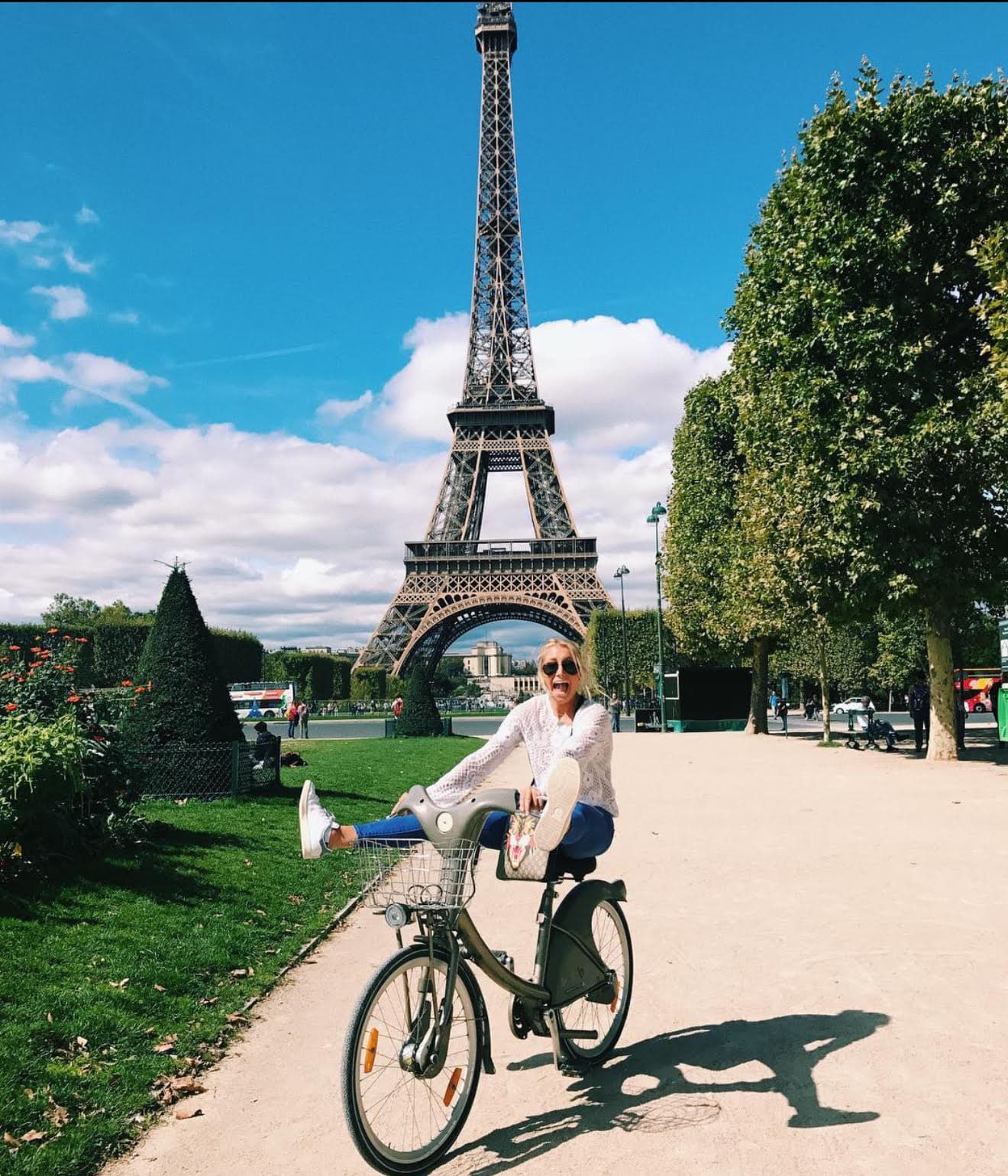 Advisor on a bicycle kicking her legs up and smiling while riding in front of the Eiffel Tower on a sunny day.