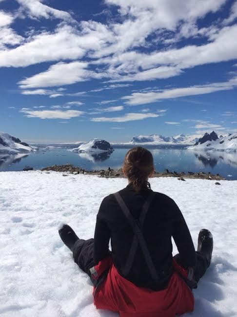 Advisor sitting in the snow facing a glassy ocean and large ice bergs on a partly cloudy day.
