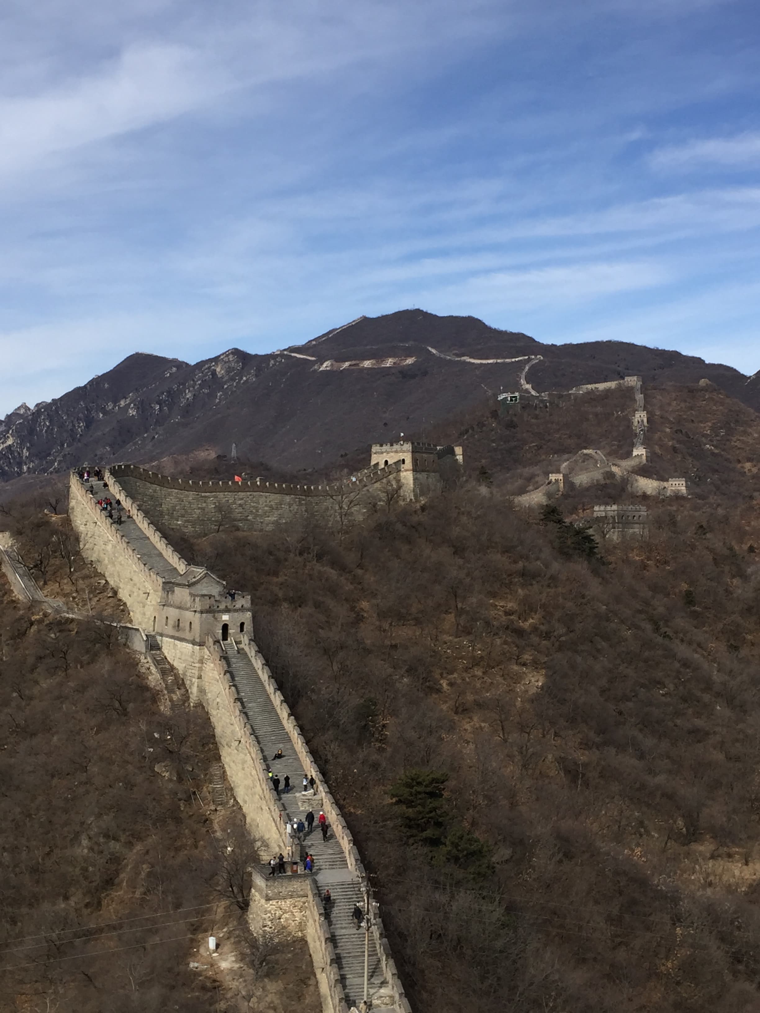 A view of a large stone wall crossing a mountain range with people walking along it on a partly cloudy day.