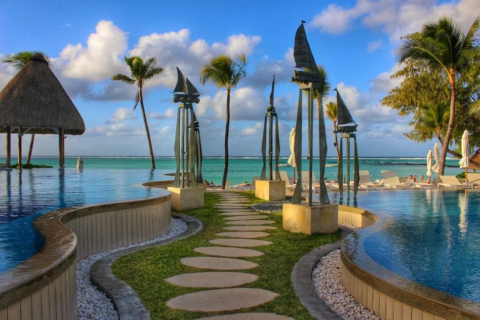 A view of a tropical beachfront with a grassy path and stepping stones leading towards the ocean with a pool on either side, a cabana and palm trees on a partly cloudy day.