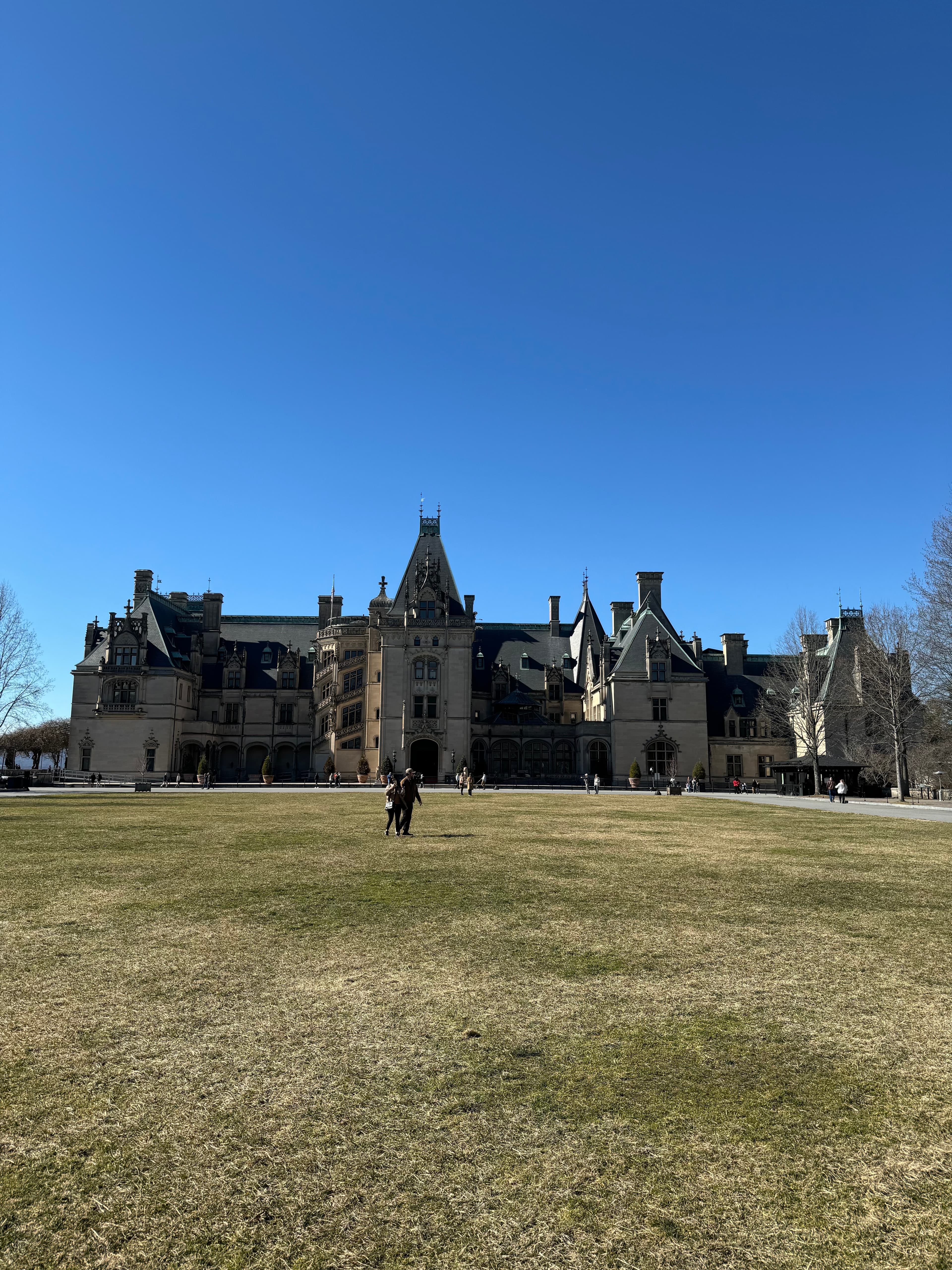 A large historical building with ornate architecture and tourists walking in a grassy lawn in front of the building.