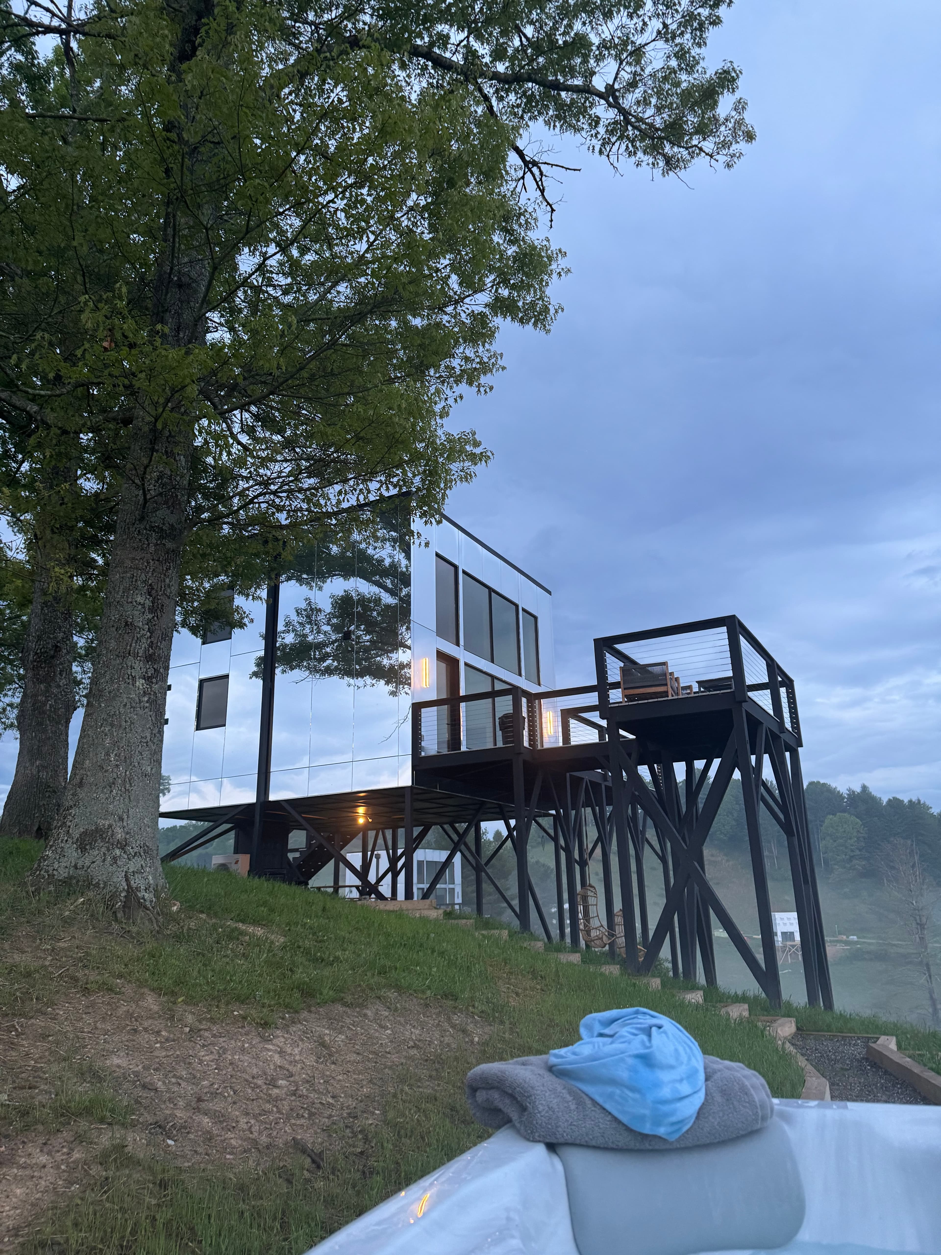 A view looking at a home with mirrored siding on a hill with a deck on stilts on an overcast day.