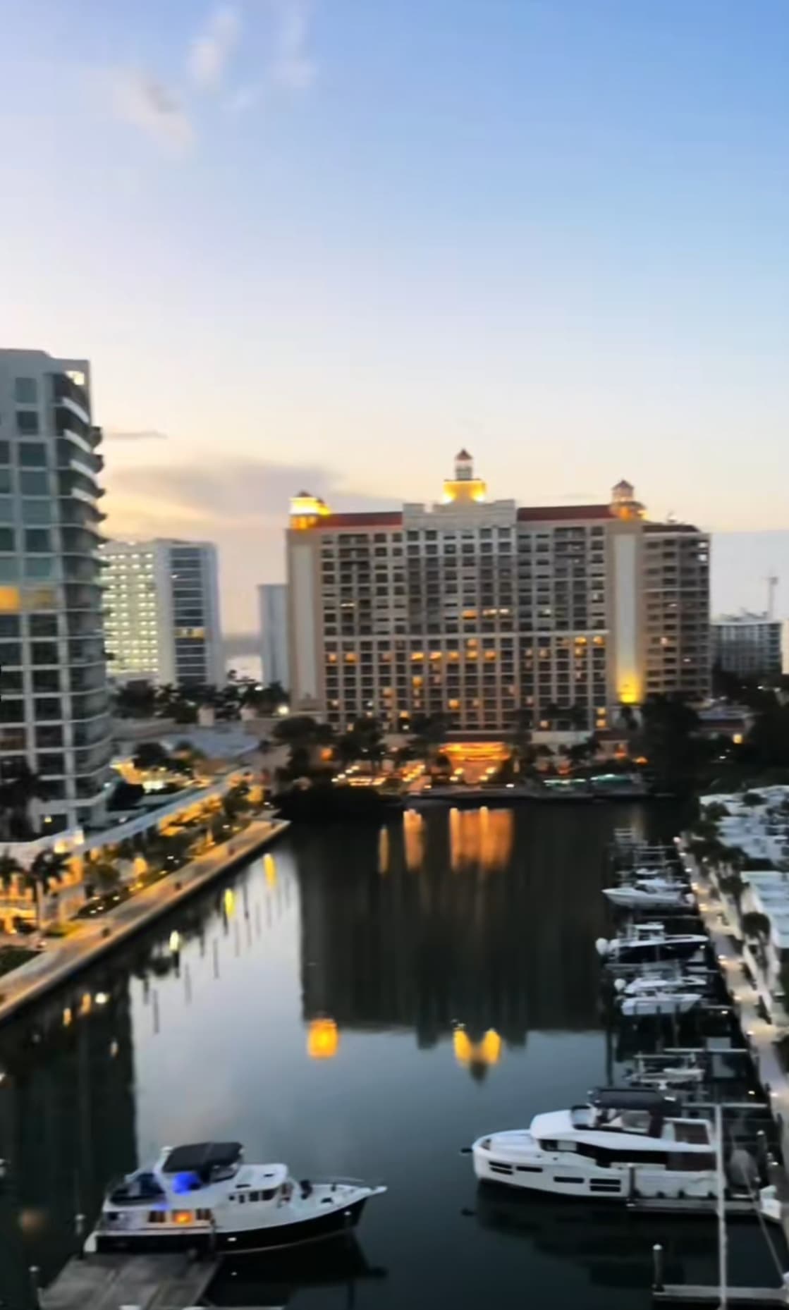 A view of a square body of water surrounded by city buildings and boats docked at sunset.