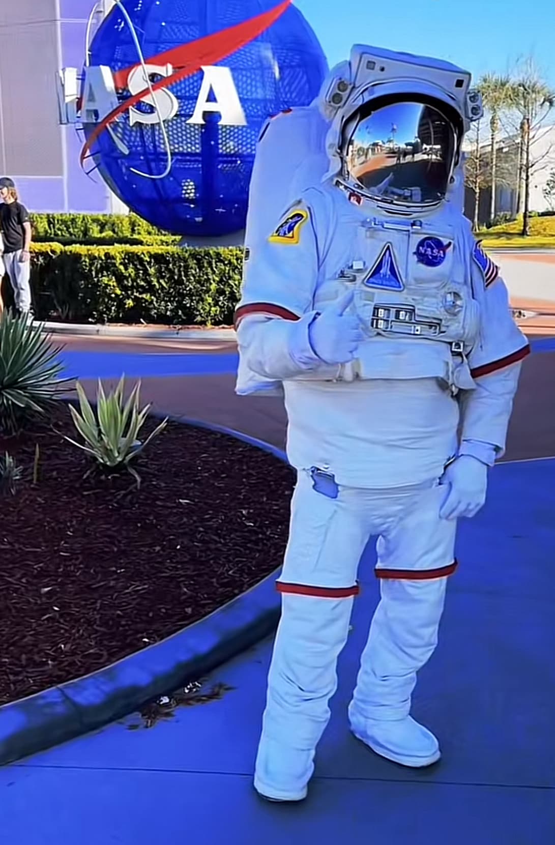 A person in an astronaut suit in posing front of a blue Nasa sign during the day.