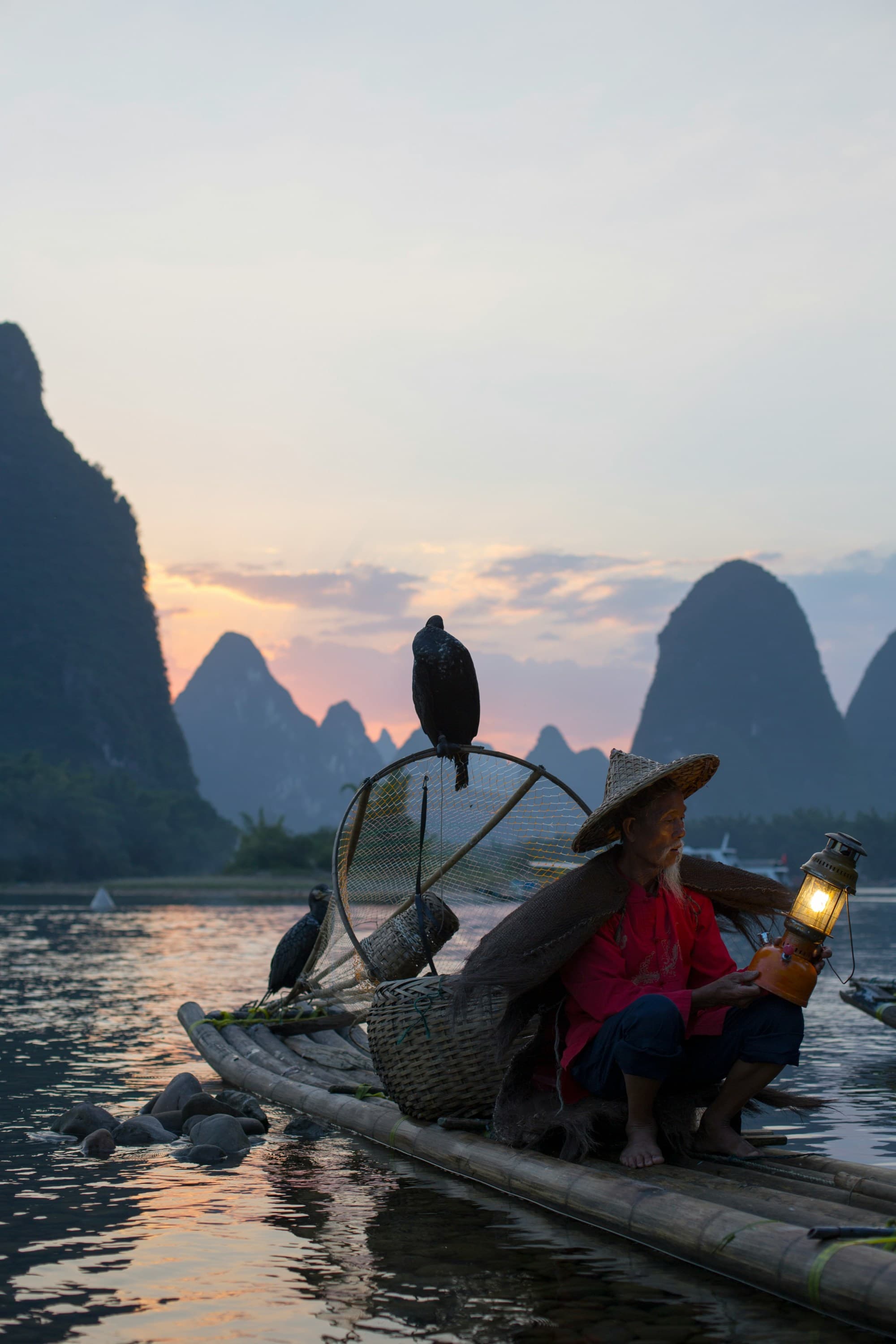 A man sits on a small handmade raft with a lantern at sunset, two birds are perched on the raft with him and there are large rock formations in the ocean ahead of them.