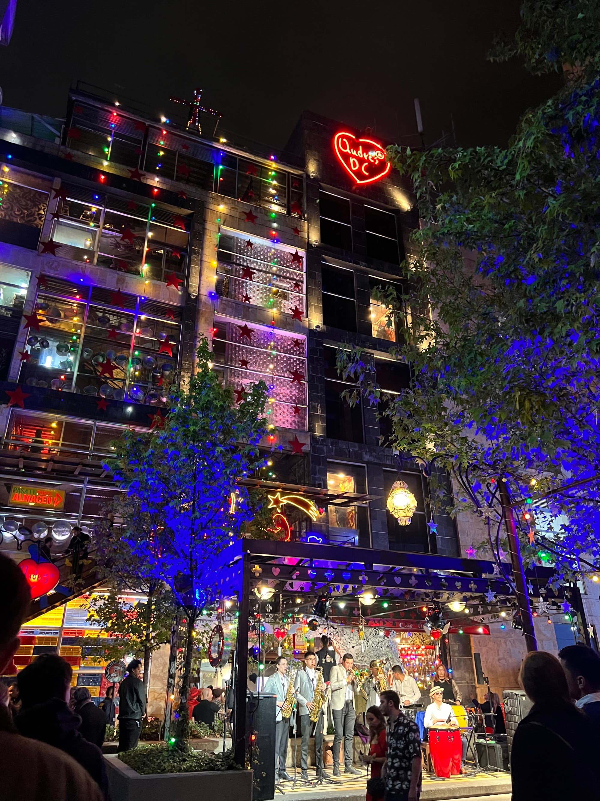 The outside of a tall city building lit up at nighttime with a large group of people on the ground level