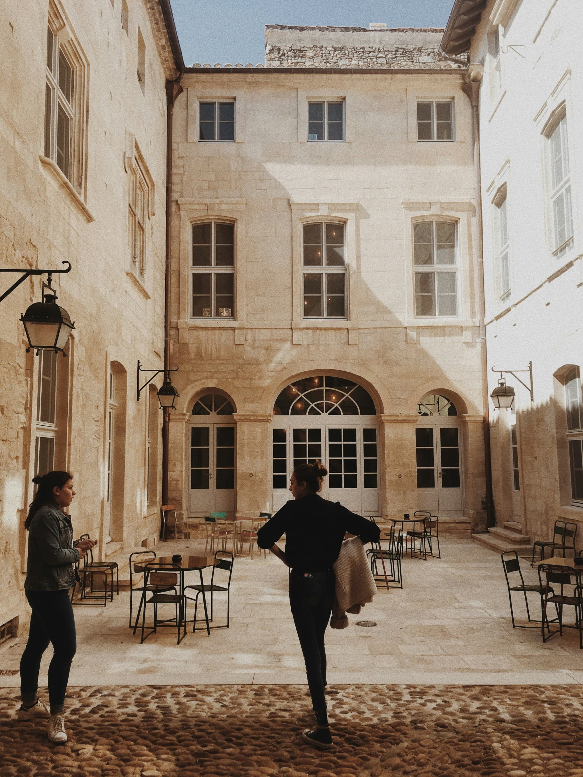 A sunlit courtyard with two people and outdoor seating in front of a classic building.