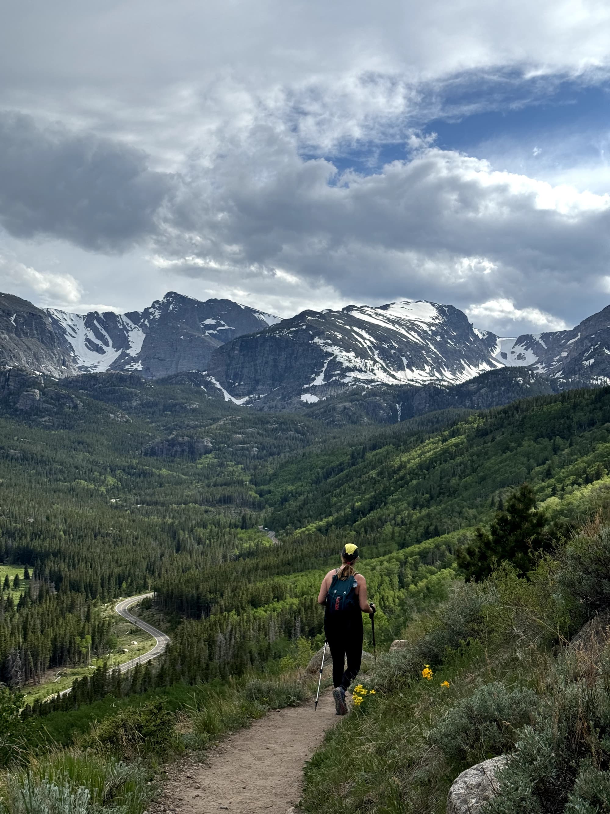 A person hikes on a trail with mountains in the background under a cloudy sky.