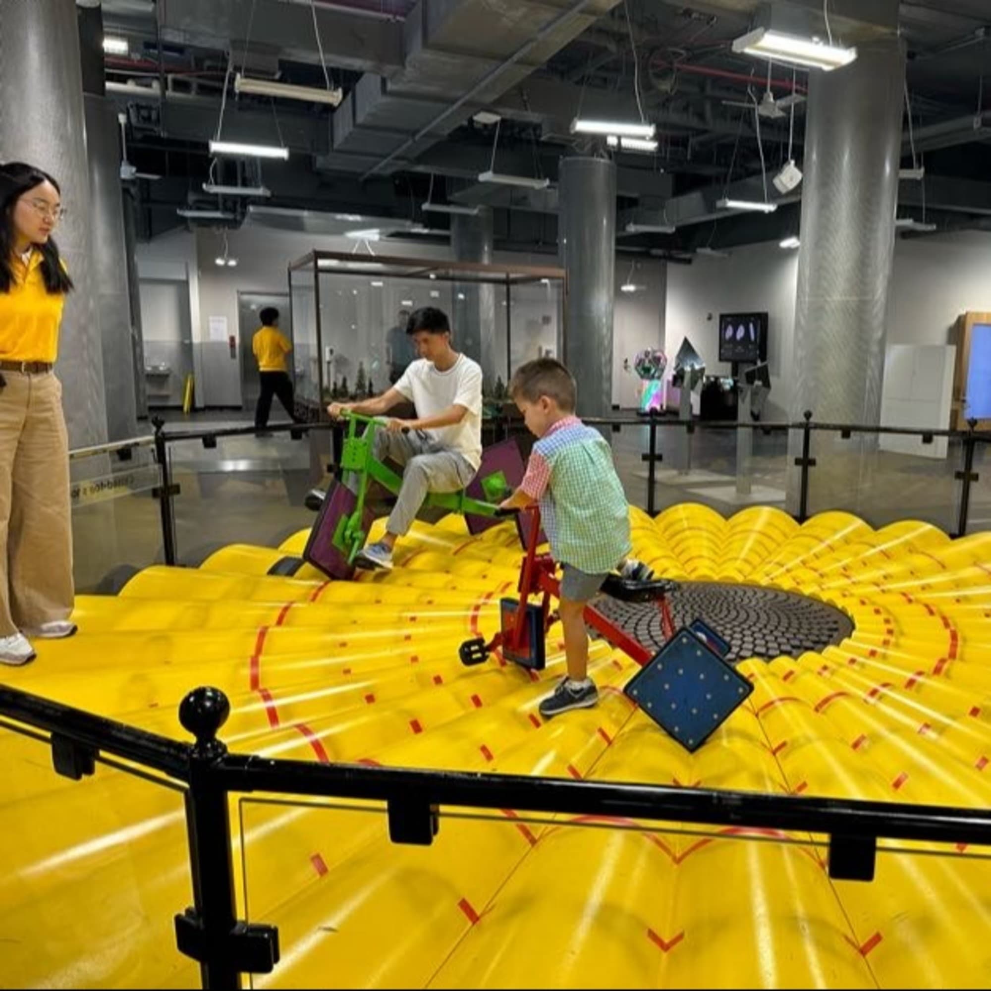 Two individuals stand on a large, yellow, circular platform with concentric circles and red arrows, surrounded by a black railing in an indoor space.
