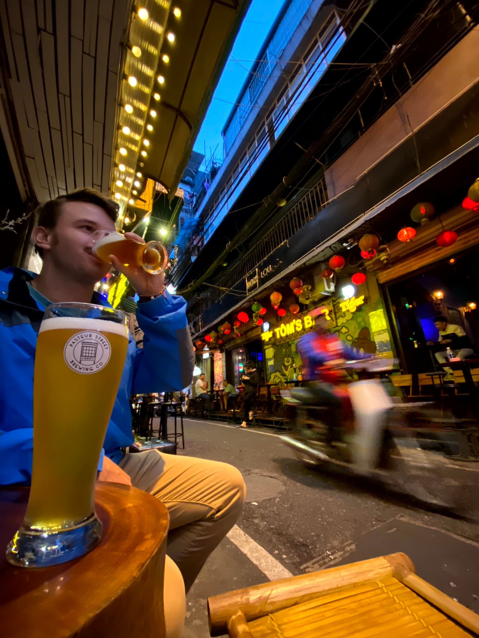 A blurred urban street scene at night, viewed from above, with a focus on a person’s hand holding a beverage.