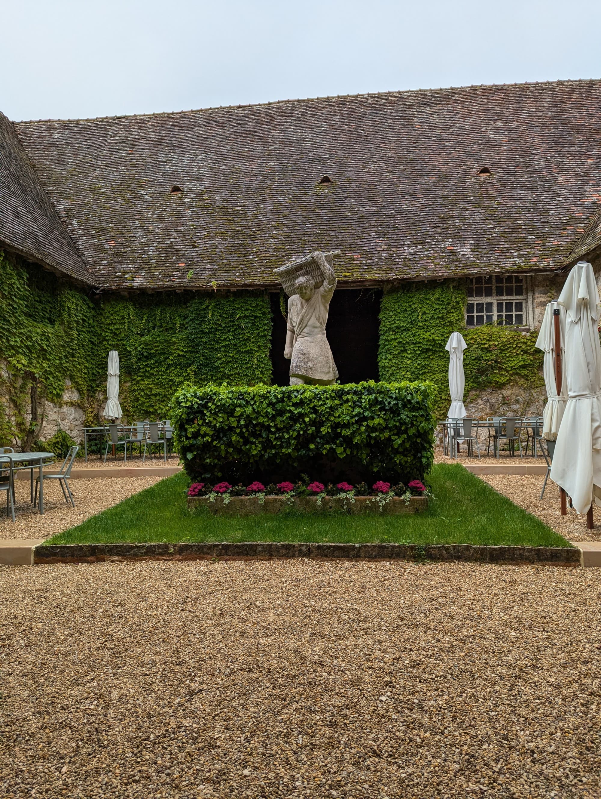 A serene garden courtyard with gravel ground, green hedges, sculptures and outdoor furniture.