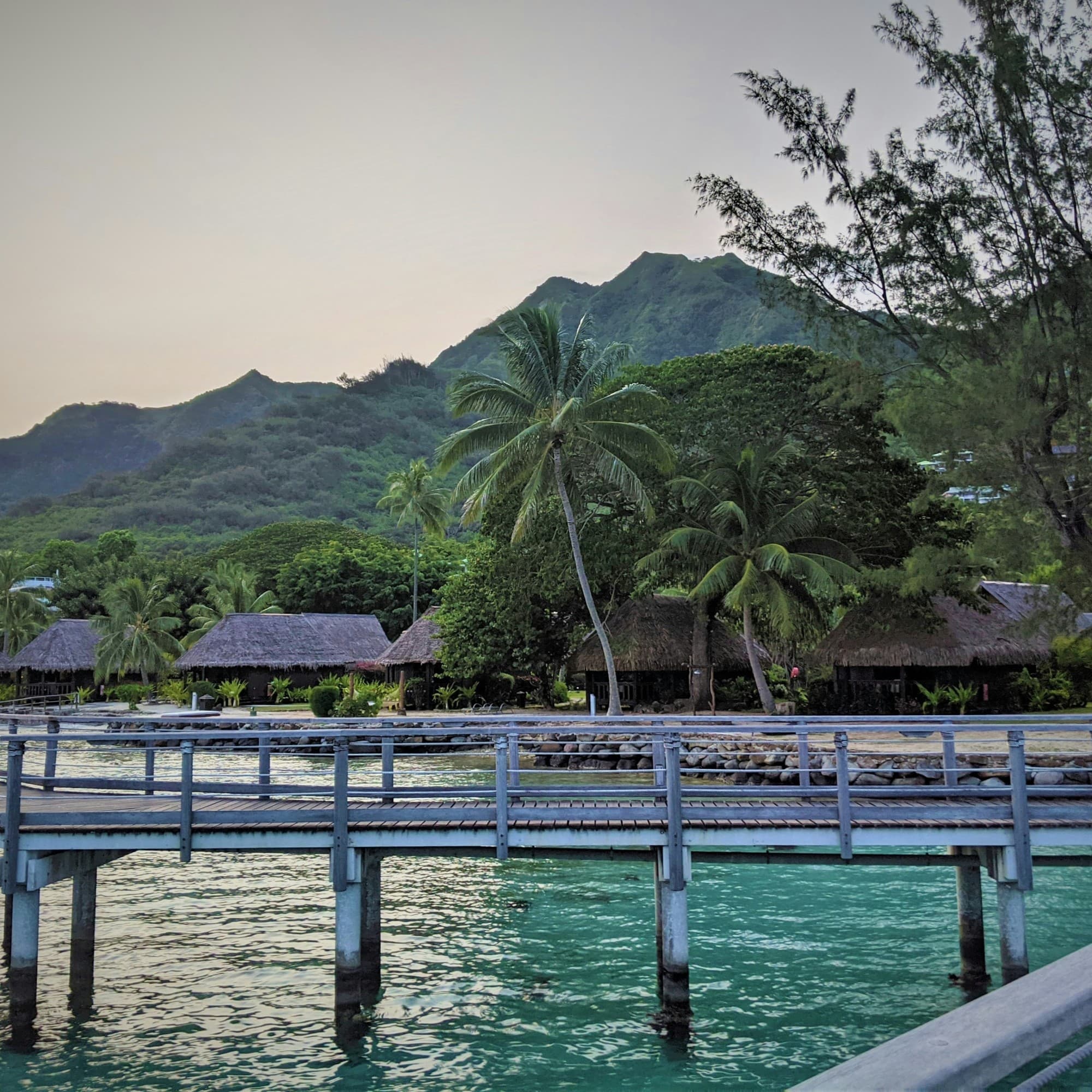 A view of a pier over clear blue water and beachside bungalows in Moorea with palm trees and a mountain in the background at sunset.