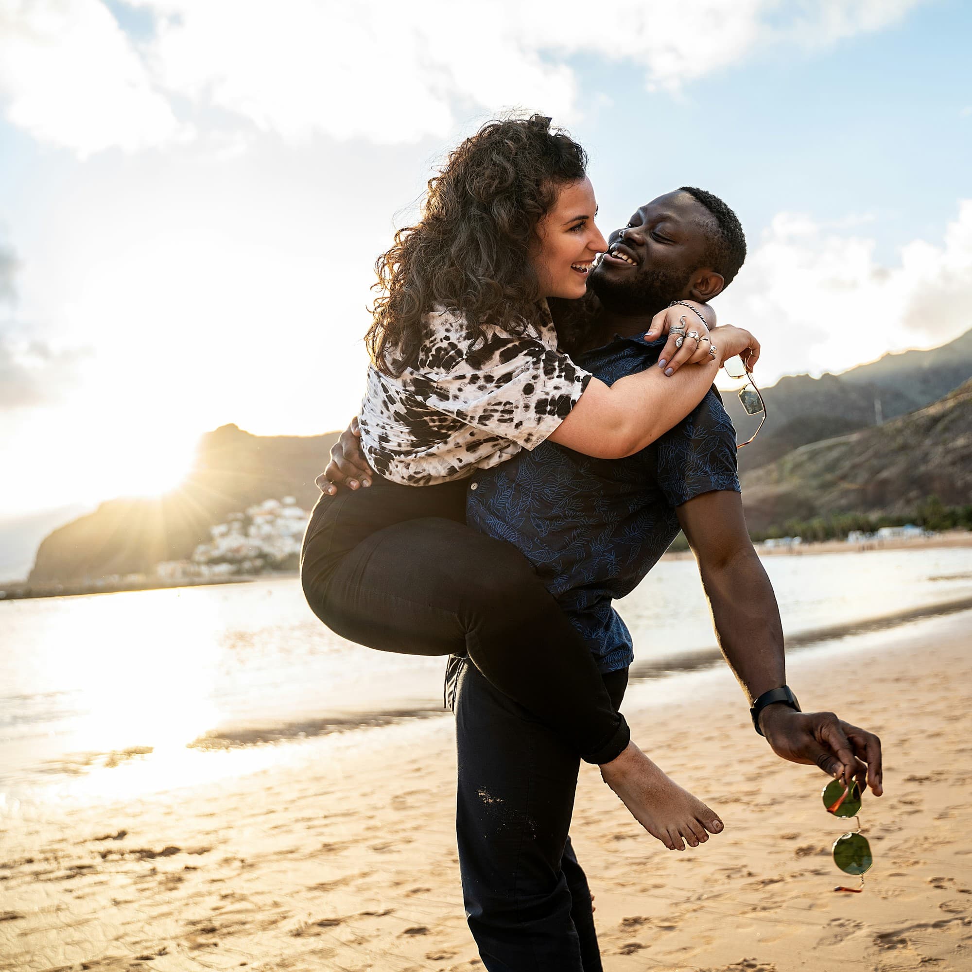 The image depicts two individuals on a beach, with one carrying the other on their back, facing a mountainous landscape during what appears to be sunset.