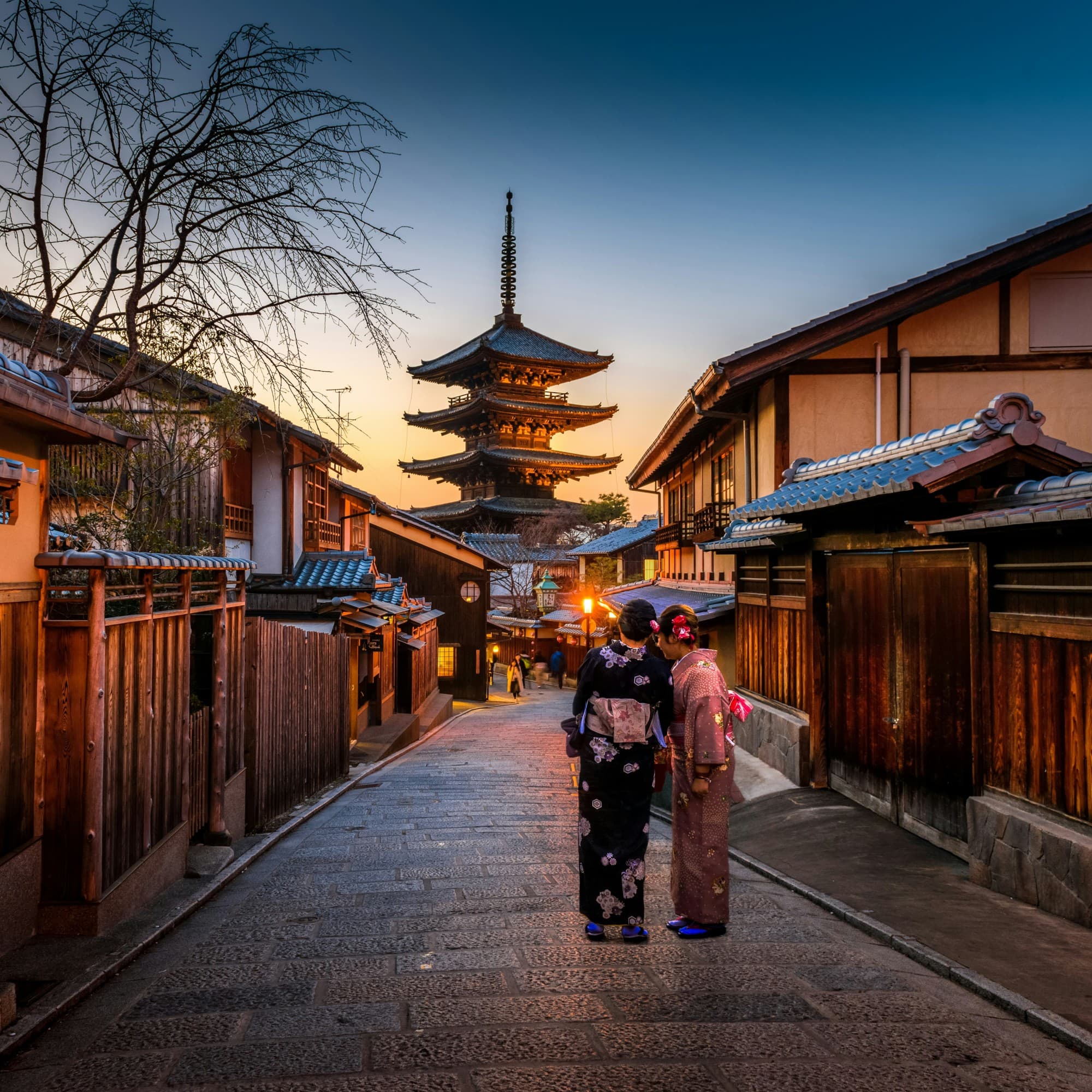 A traditional Japanese street at dusk features two individuals in kimonos with a five-story pagoda in the background.