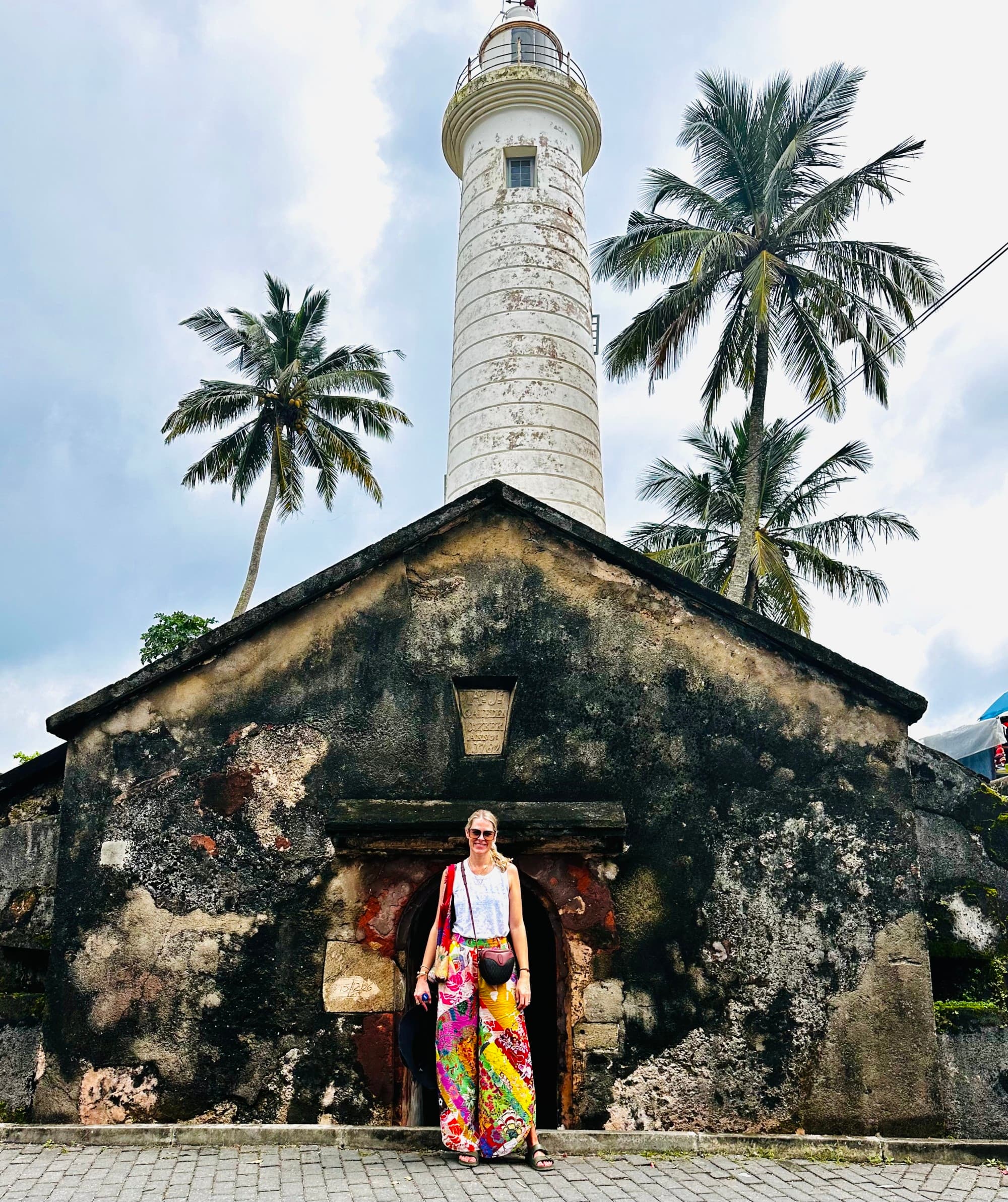 A picture of a person standing in front of an old lighthouse with palm trees under a cloudy sky.