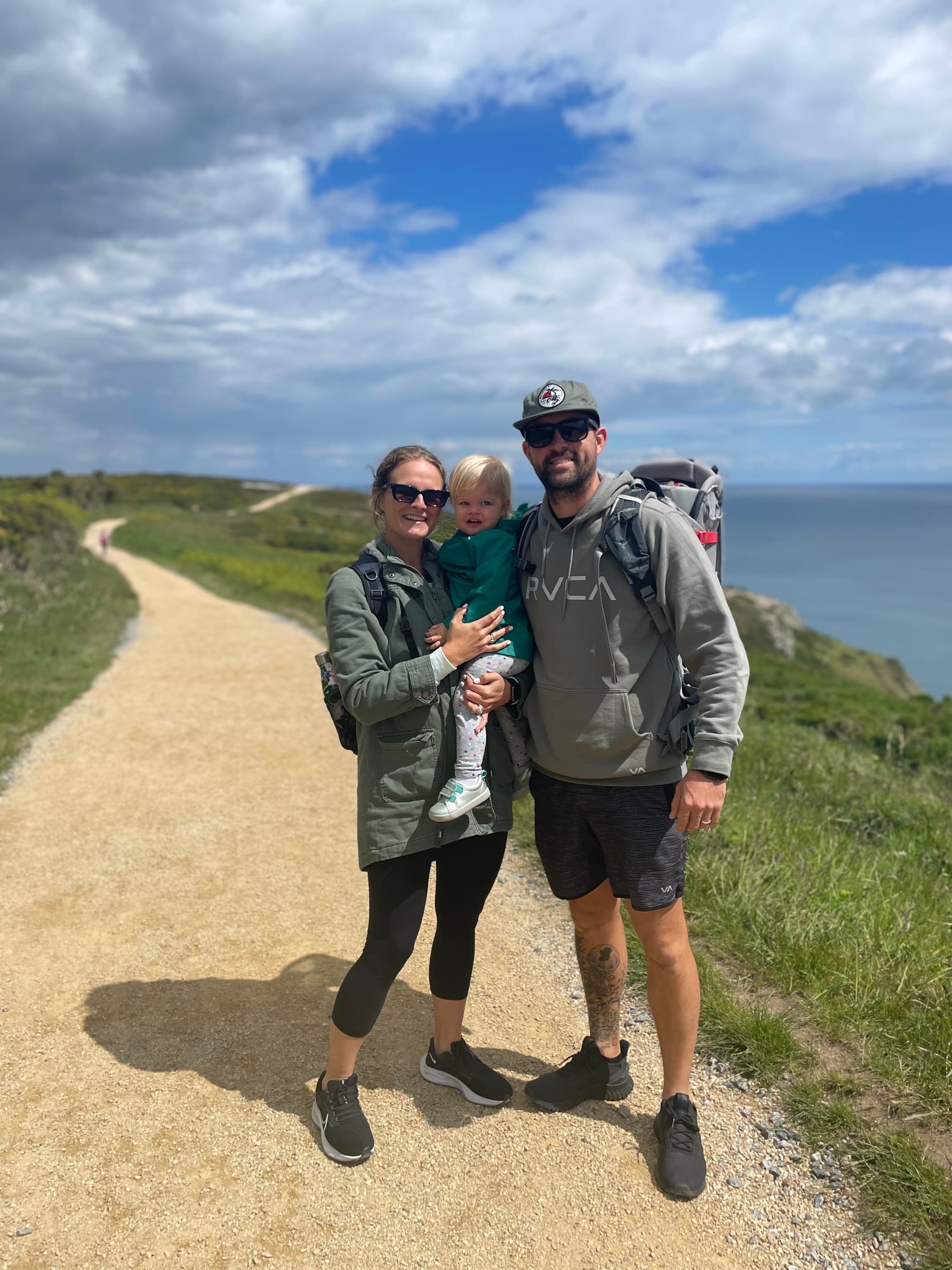 The image shows two adults and a child standing on a dirt path with a scenic ocean view in the background.