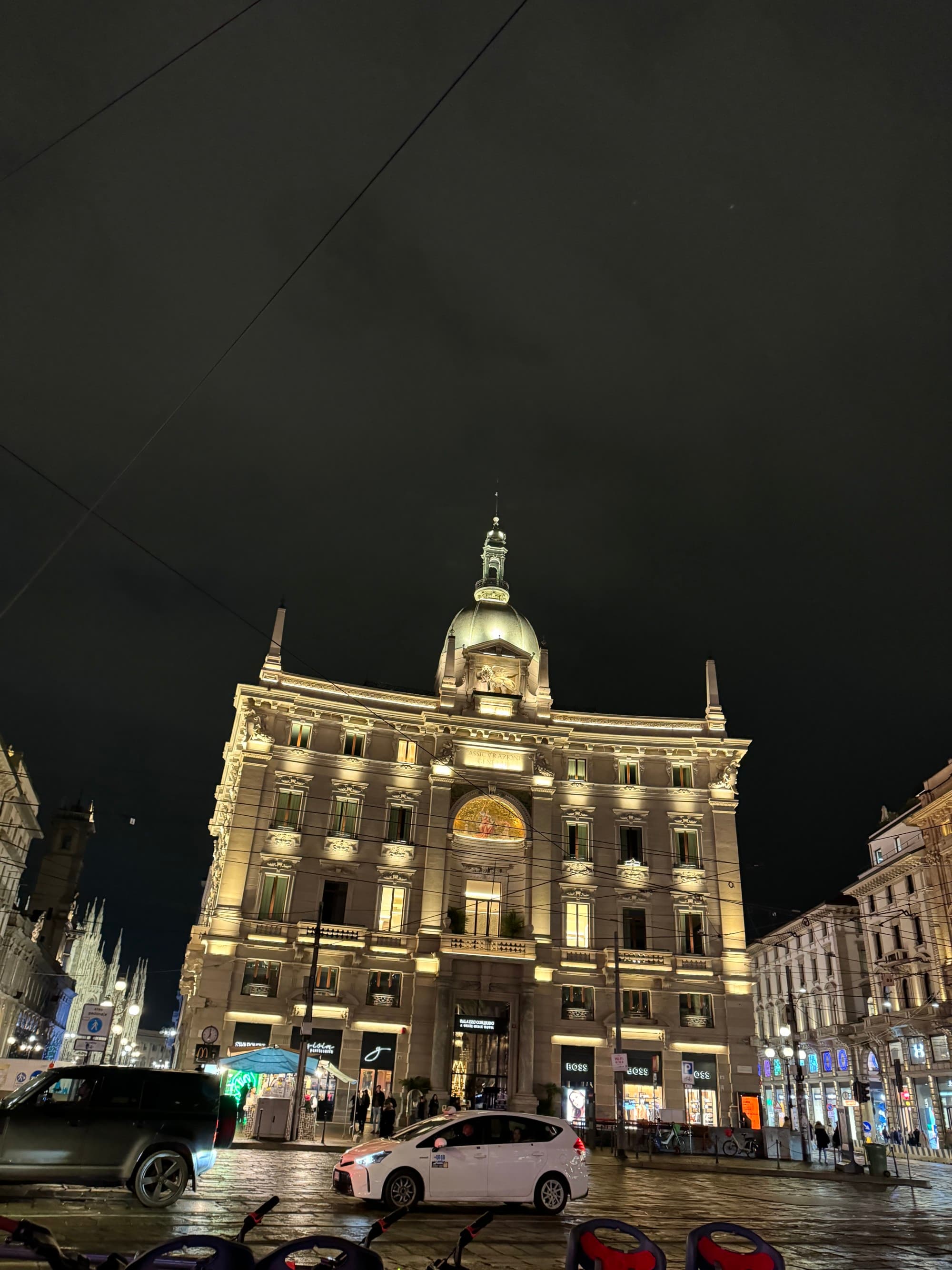 A low-angled shot of a beige-colored building near a street at night. There are cars driving in front of it.