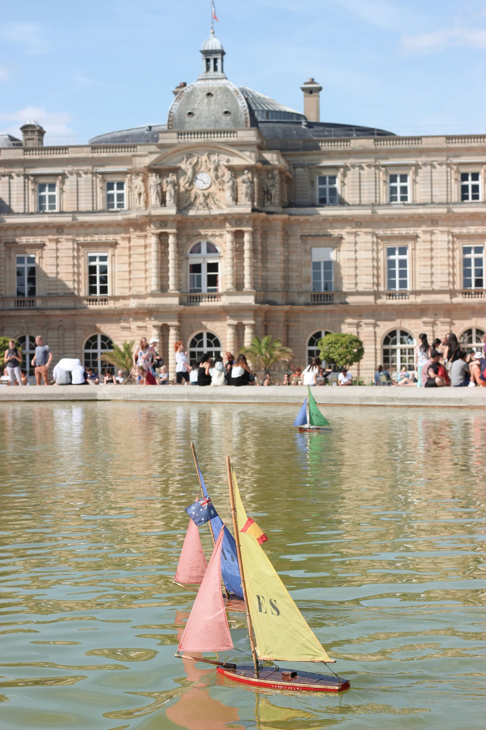 Vibrant model sailboats glide on water, with a grand edifice backdrop and onlookers at the water’s edge.