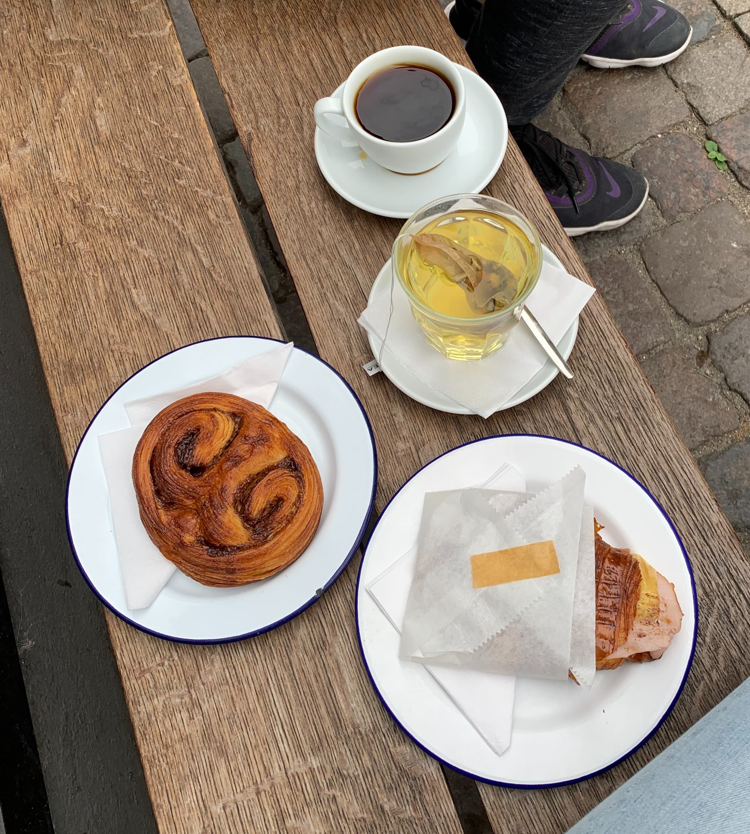 A wooden bench with four white dishes complete with coffee, tea and two pastries.