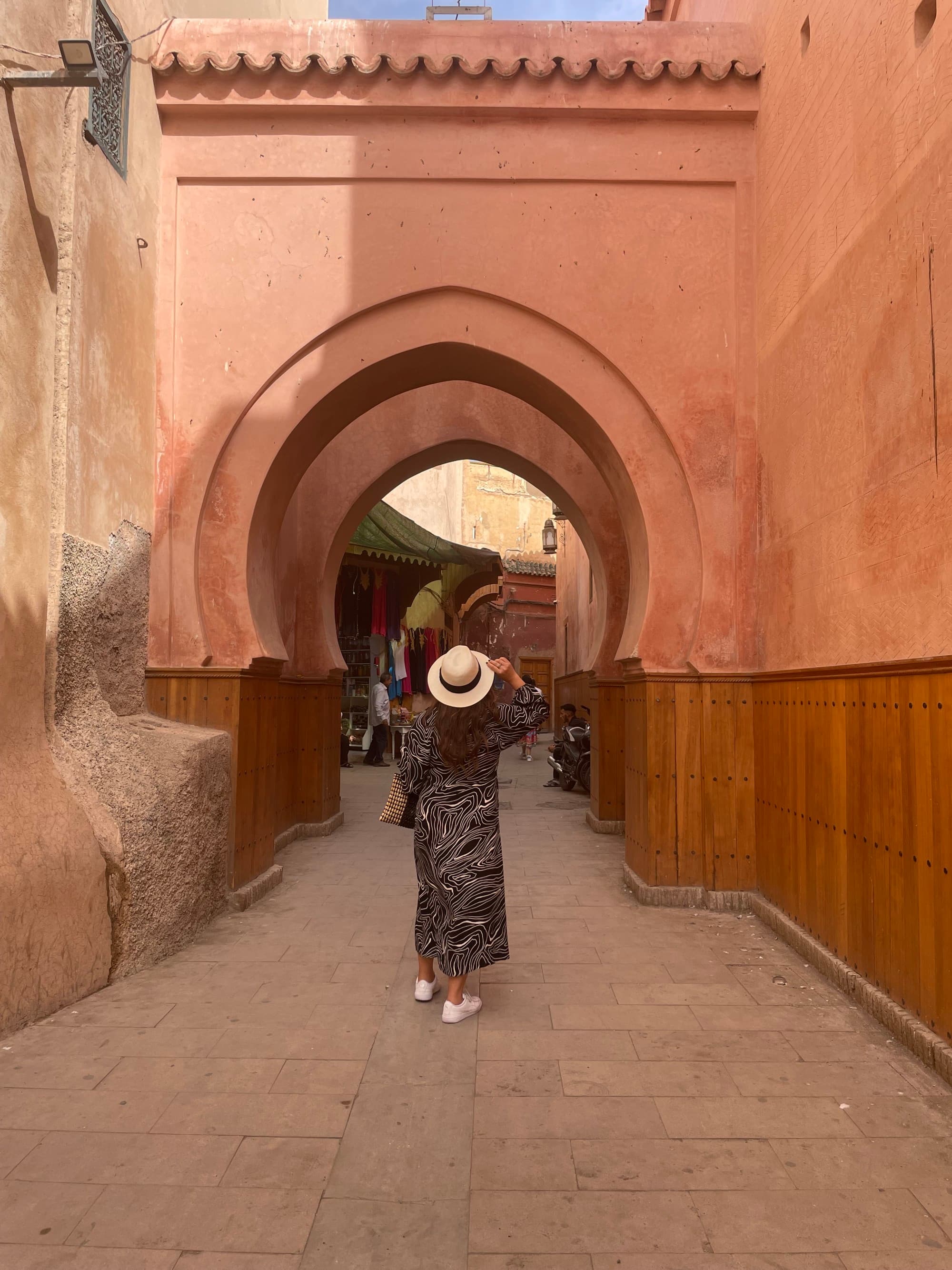 A woman wearing a white hat and a long dress posing under a brown concrete arch during daytime.