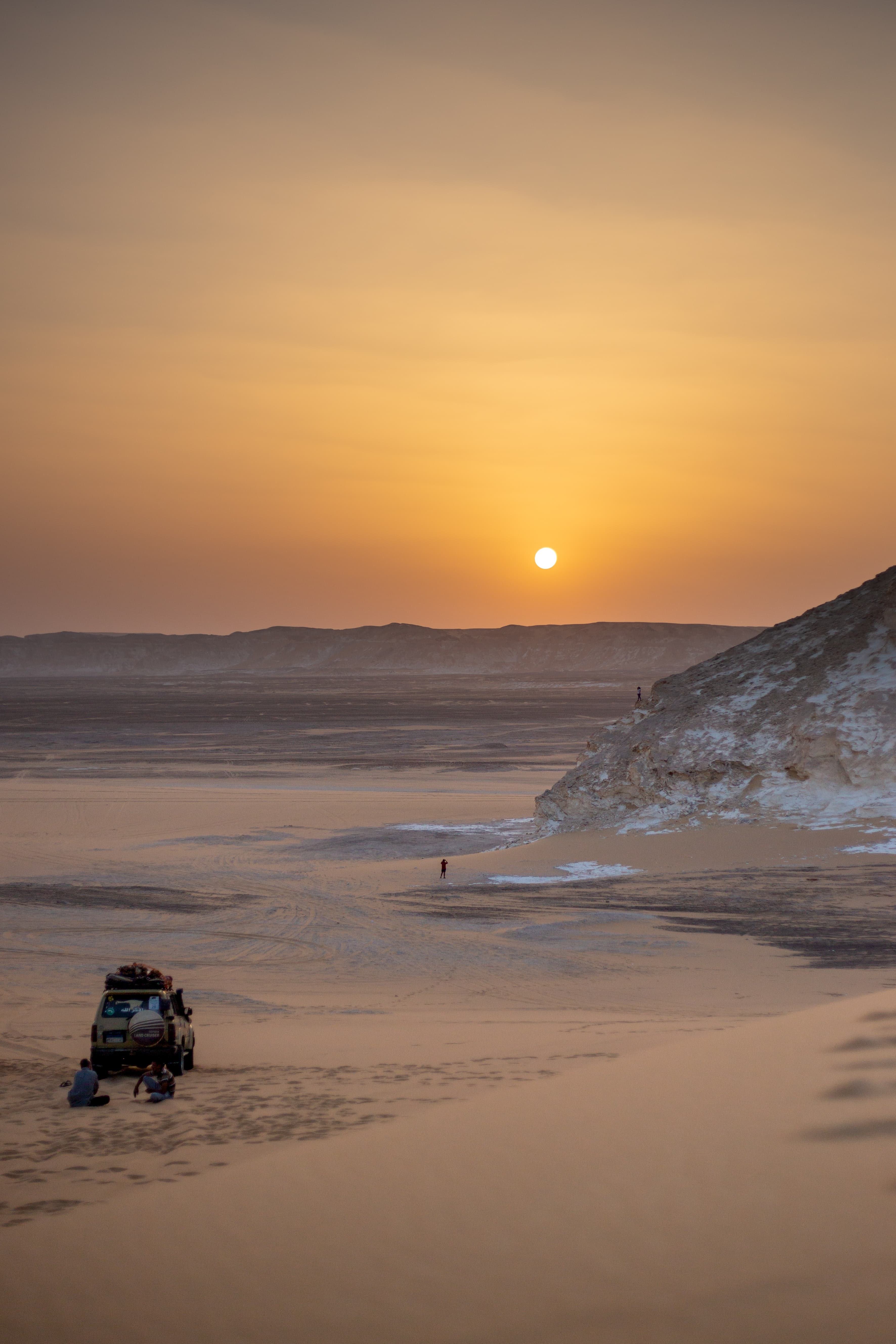 sunset over a calm desert with a car and a person walking towards the sun