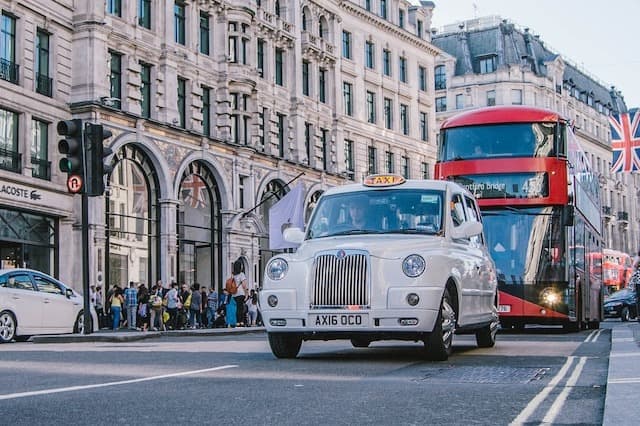A red colored double decker and a white car on the road in London.