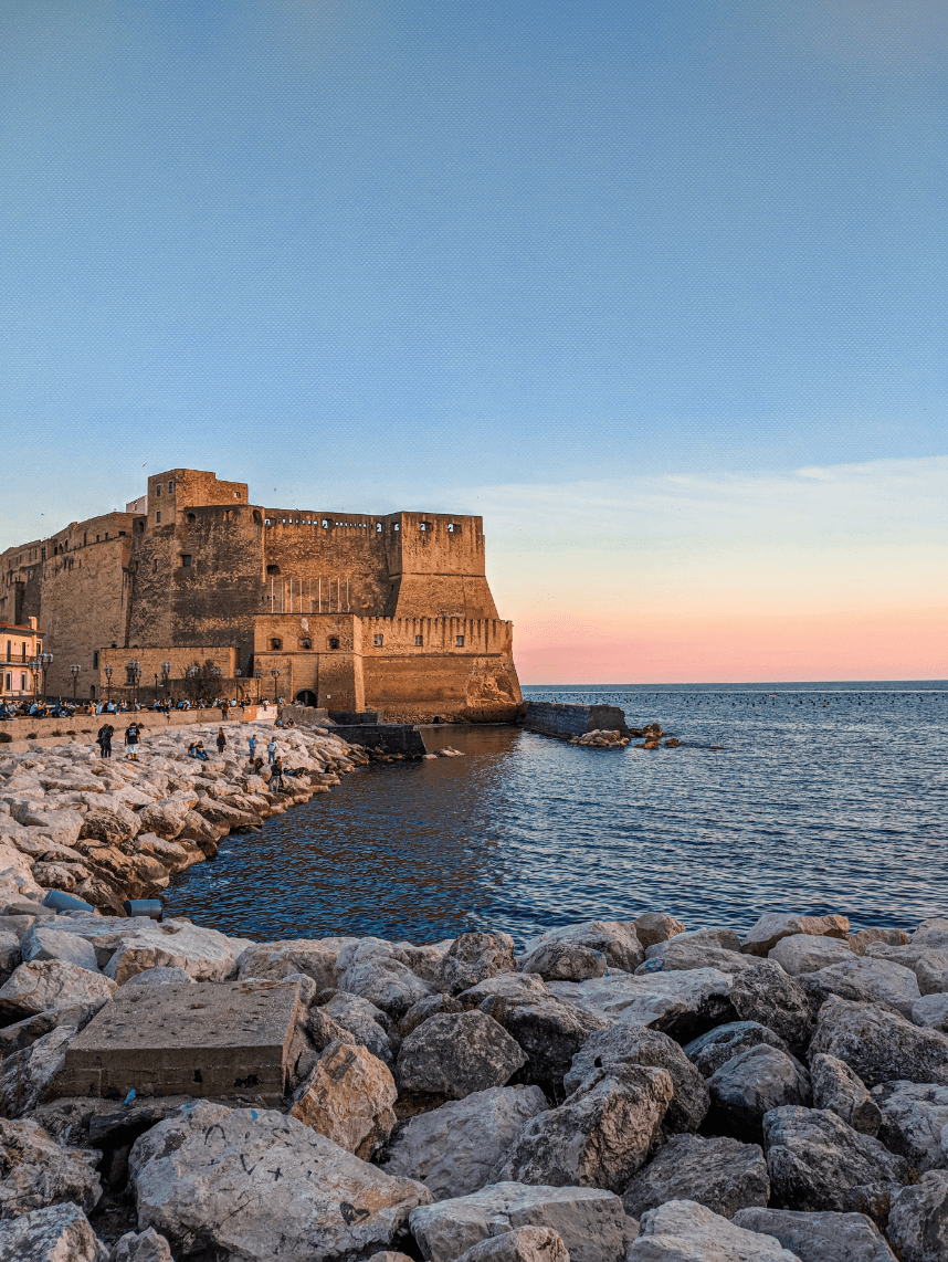 A rocky shore looking out to a rippling blue sea with a large stone fort in the background against a light pink sunset.