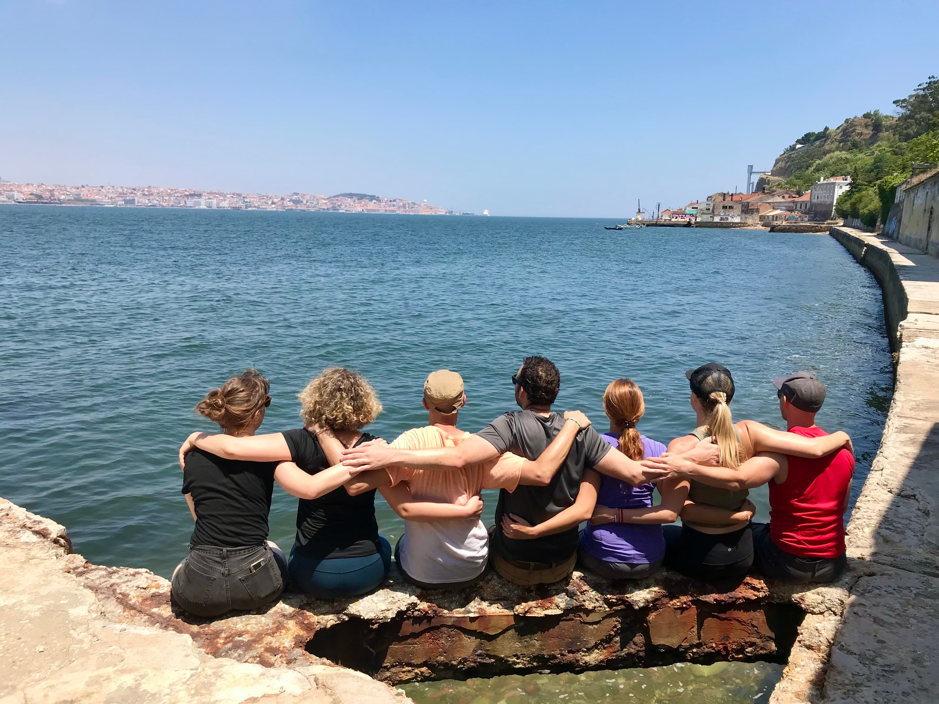 A group of people sitting on the waters edge with arms around each other, backs to the camera