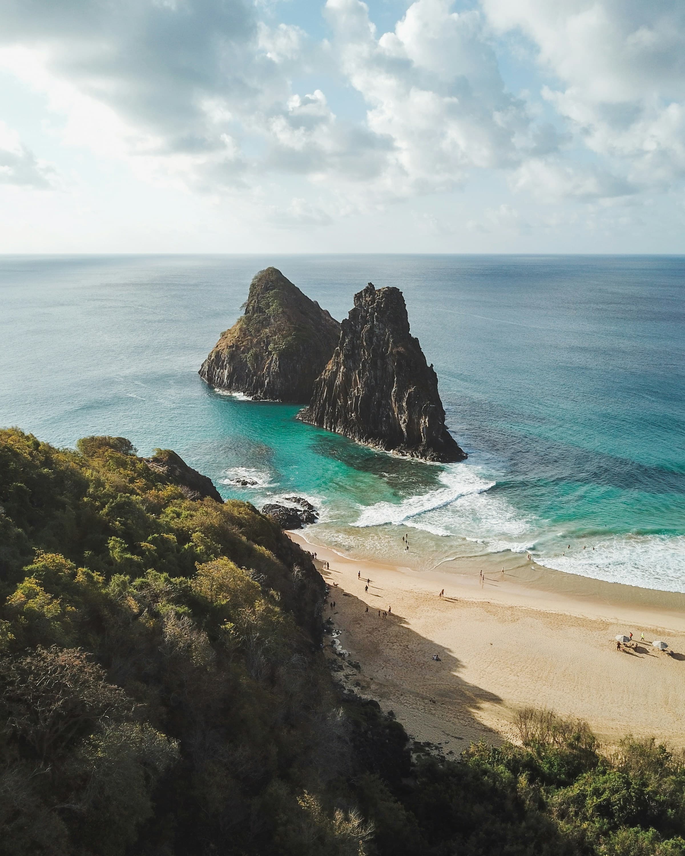 An aerial view of trees, the beach, turquoise blue water and large rock formations poking out of the water.