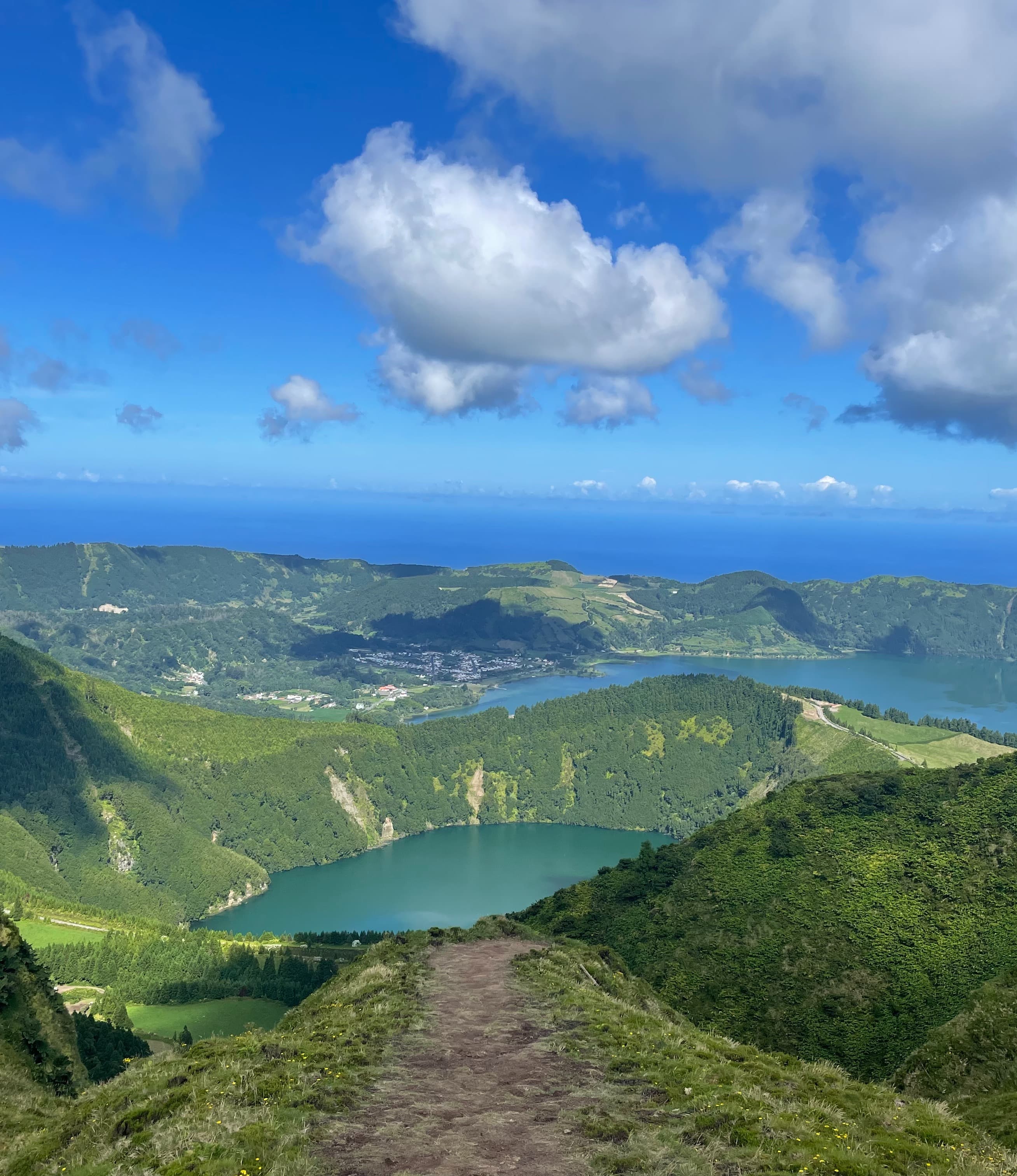 A dirt path leading to a view of a turquoise lake nestled into a green mountain range beneath the thick clouds and blue sky.