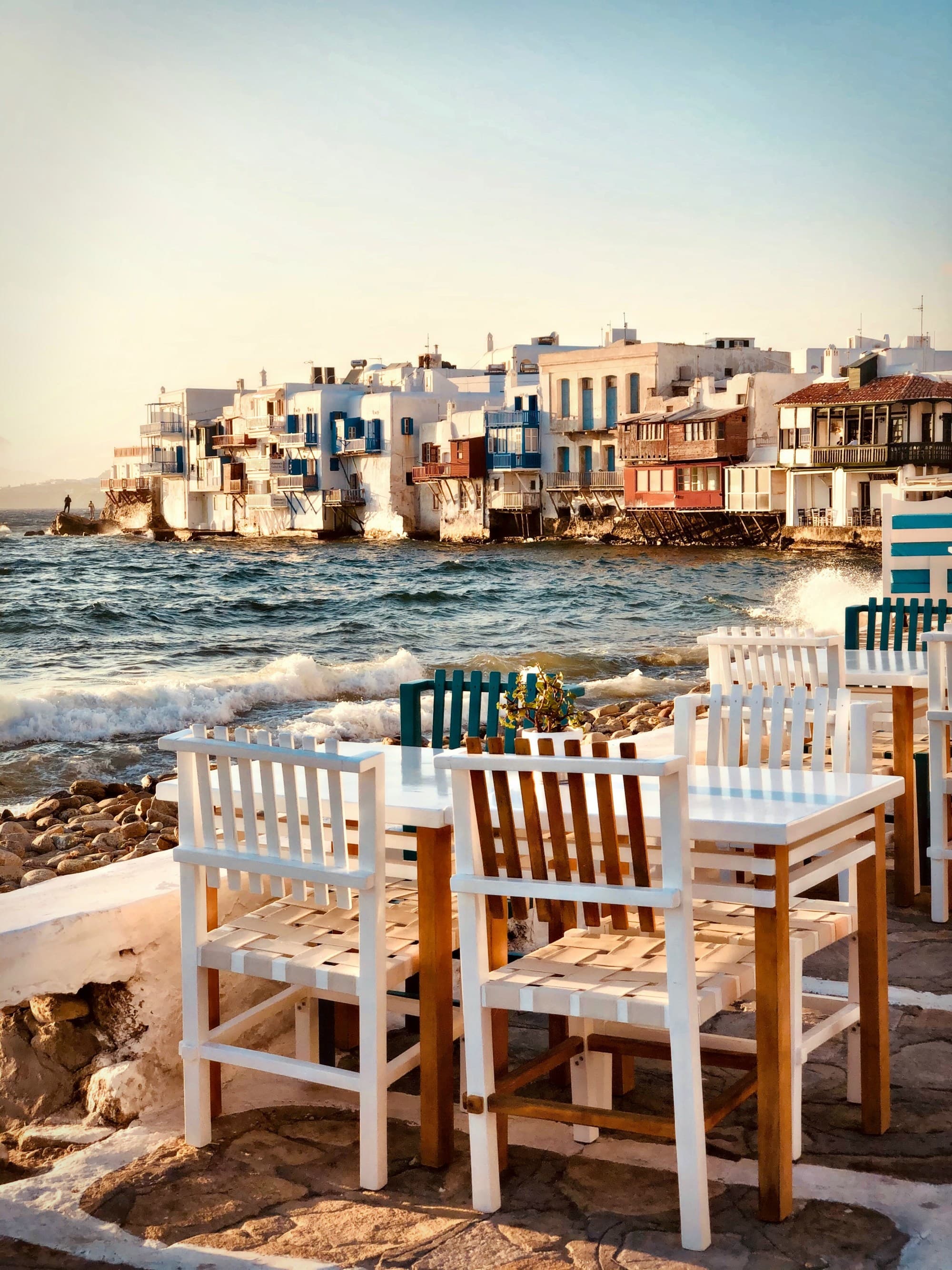 Tables and chairs near the blue water with Greek architecture in the background.