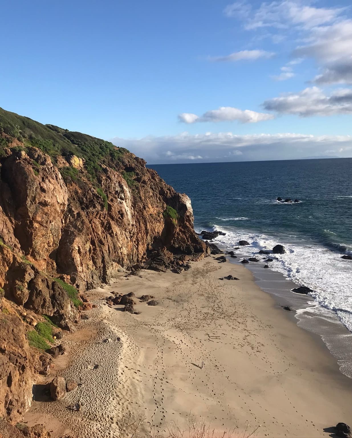A sandy beach, rocky cliff and waves crashing in from the Pacific Ocean.