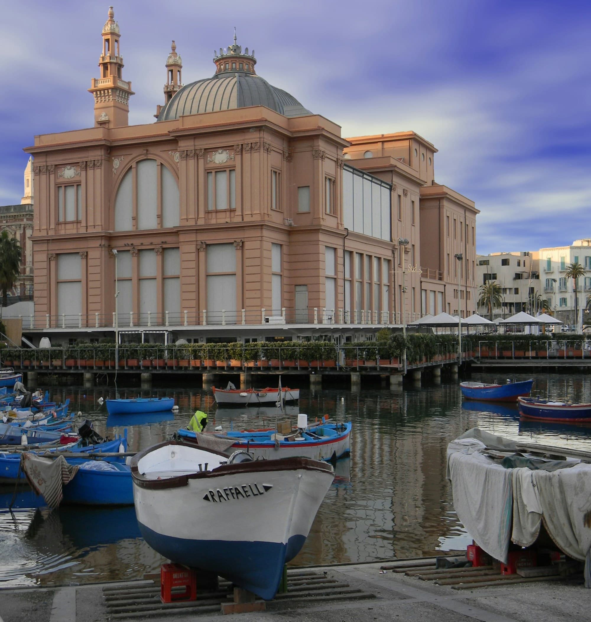 A group of boats anchored in a harbor with an elaborate, tan building in the background under a vibrantly blue and cloudy sky.