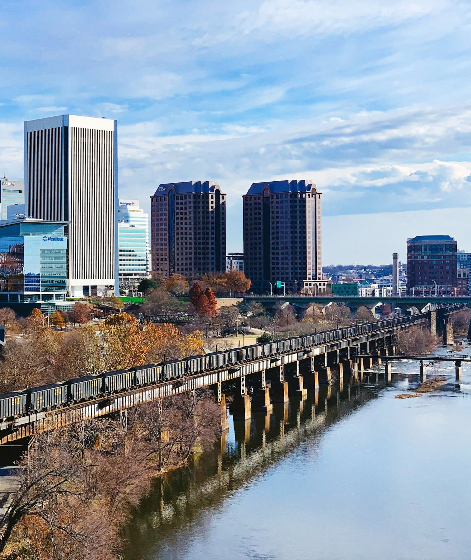 A view of Richmond, featuring a river, bridge, Autumnal trees and skyscrapers under a cloudy blue sky.