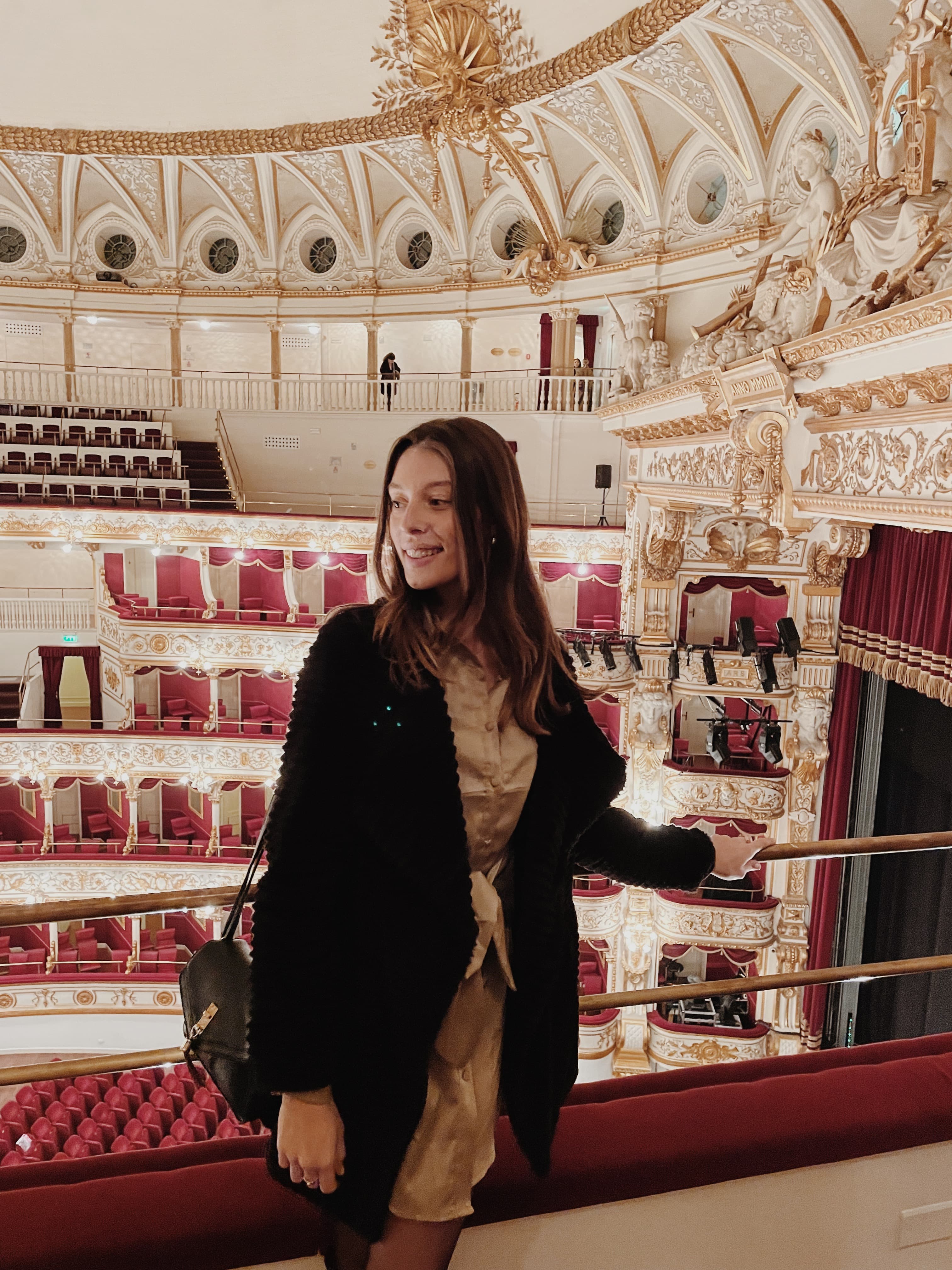 A girl posing for a photo inside of a glamorous old theatre with red seats and white and gold details.
