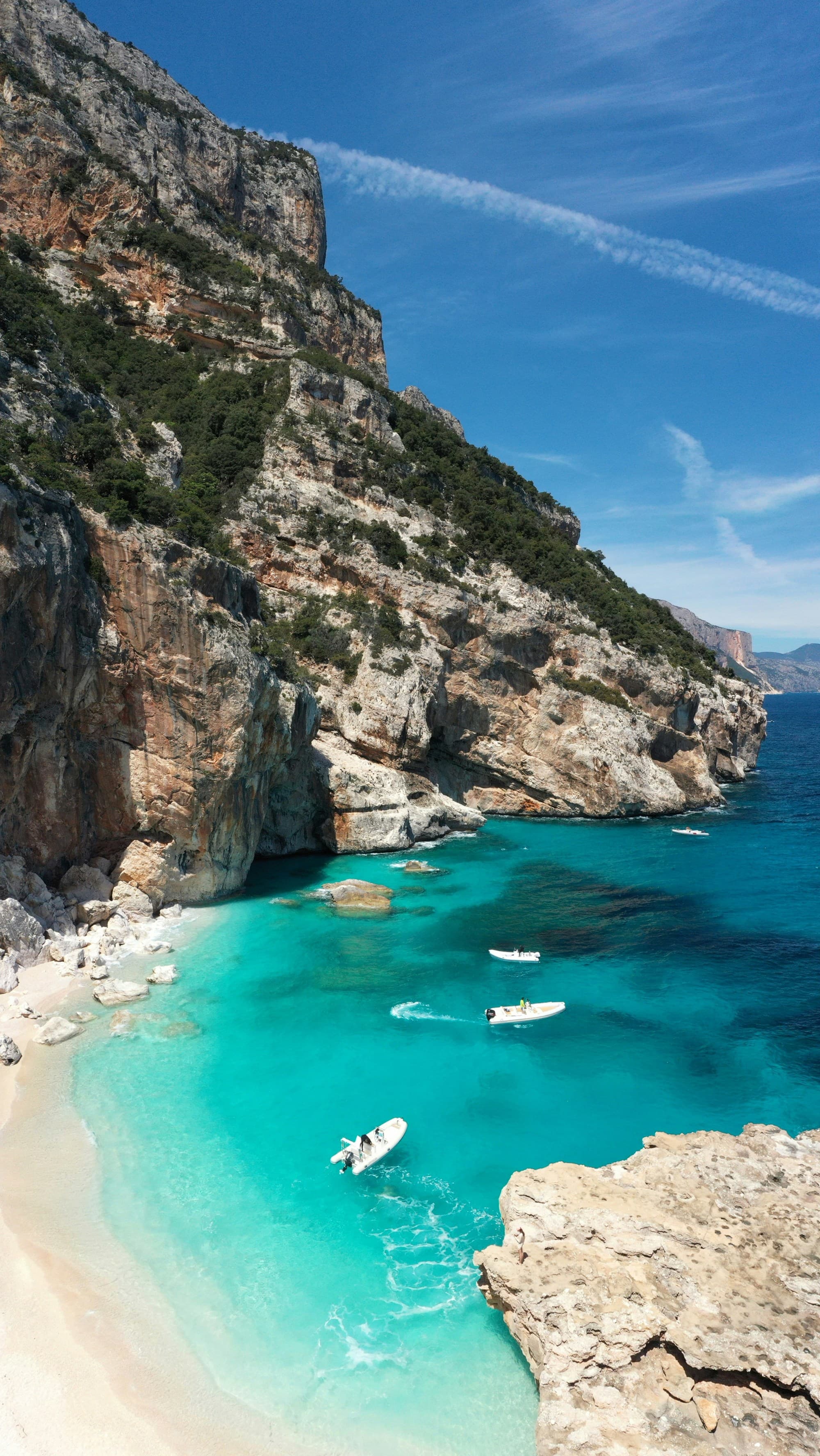 An aerial view of the vibrant beach and turquoise sea, with boats, rocks and green cliffs in the surrounding areas.