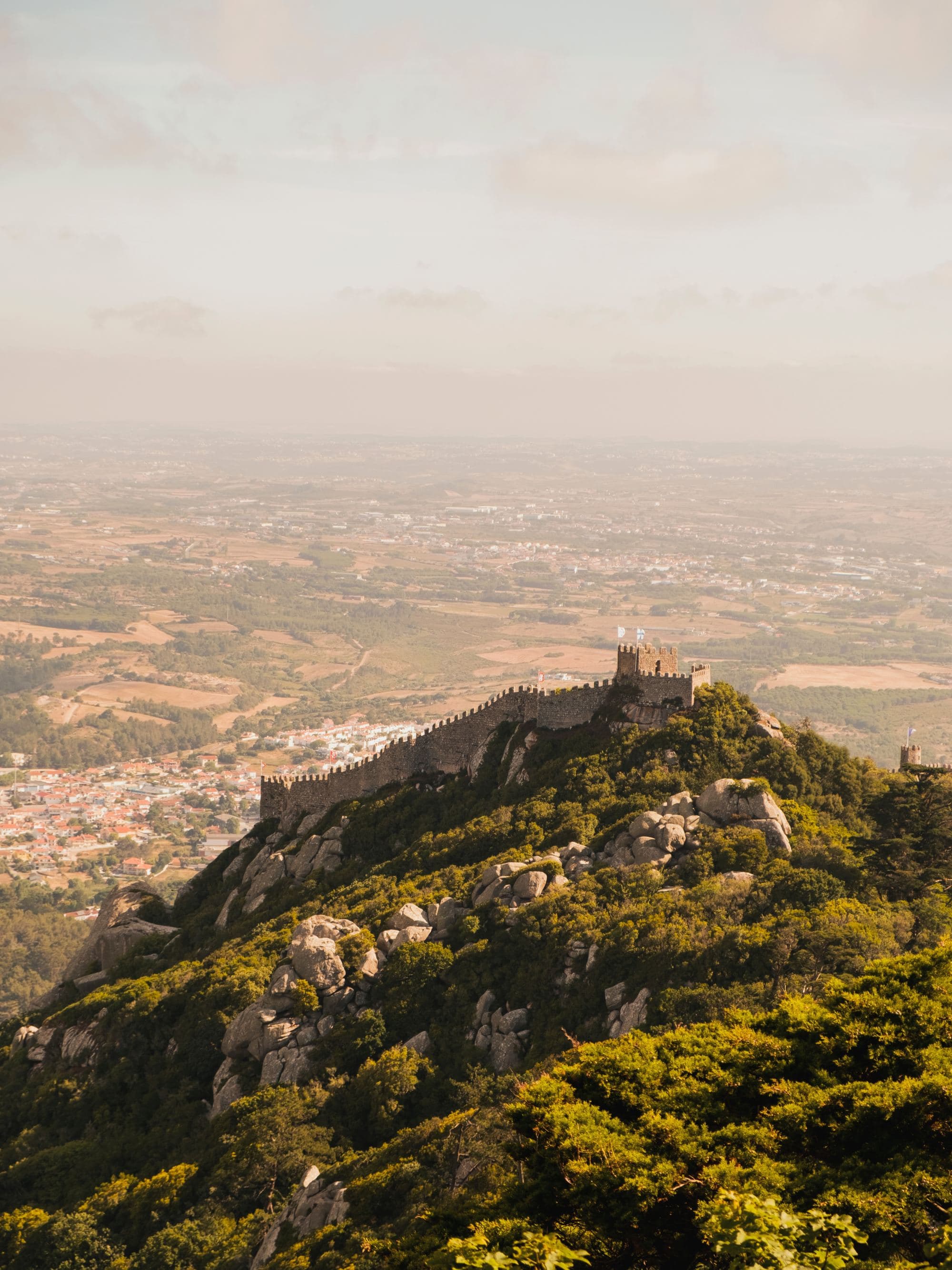 a castle a top a green hill above a countryside town on a hazy sunny day