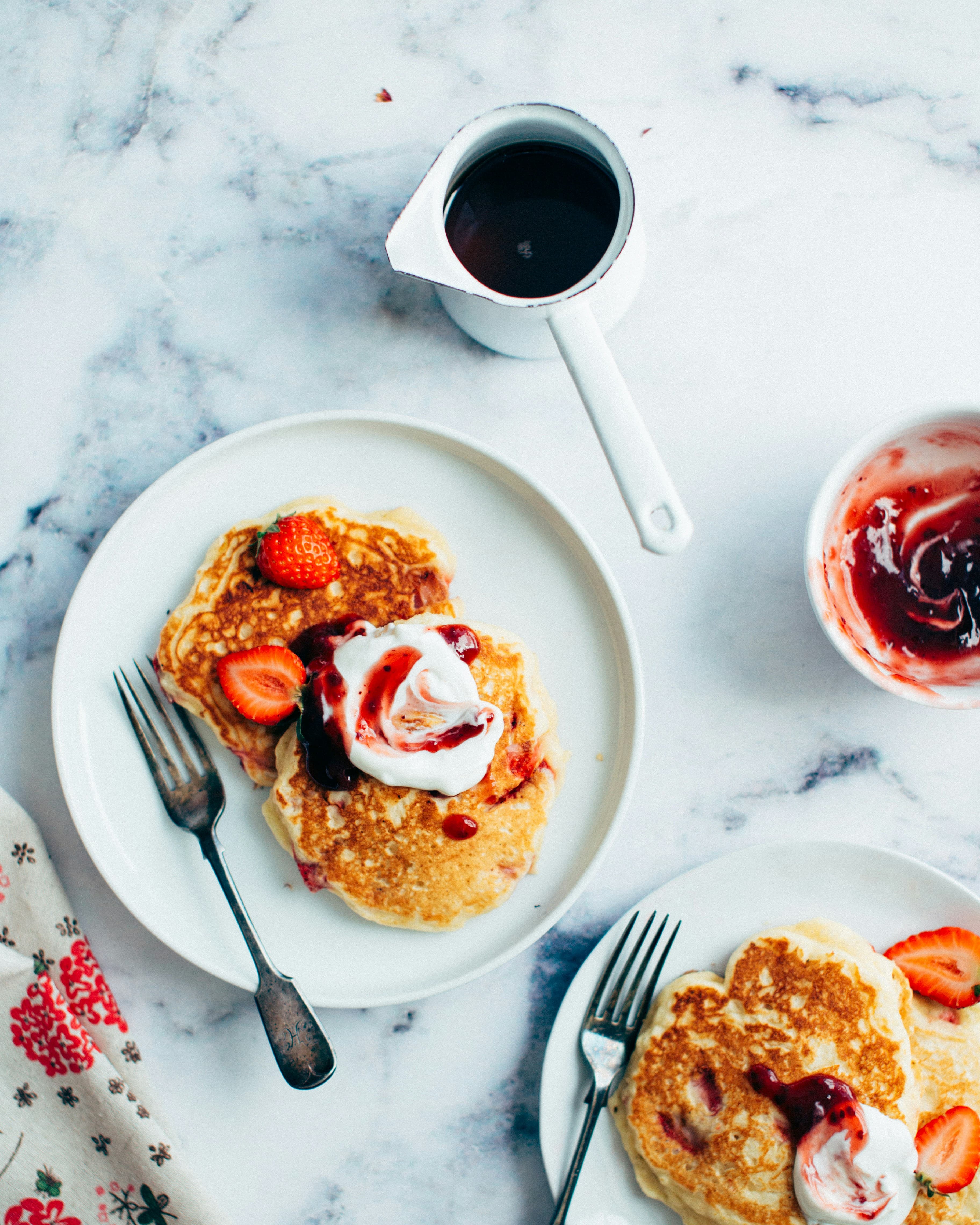 Plates of pancakes topped with whipped cream and berries on a white marble table.