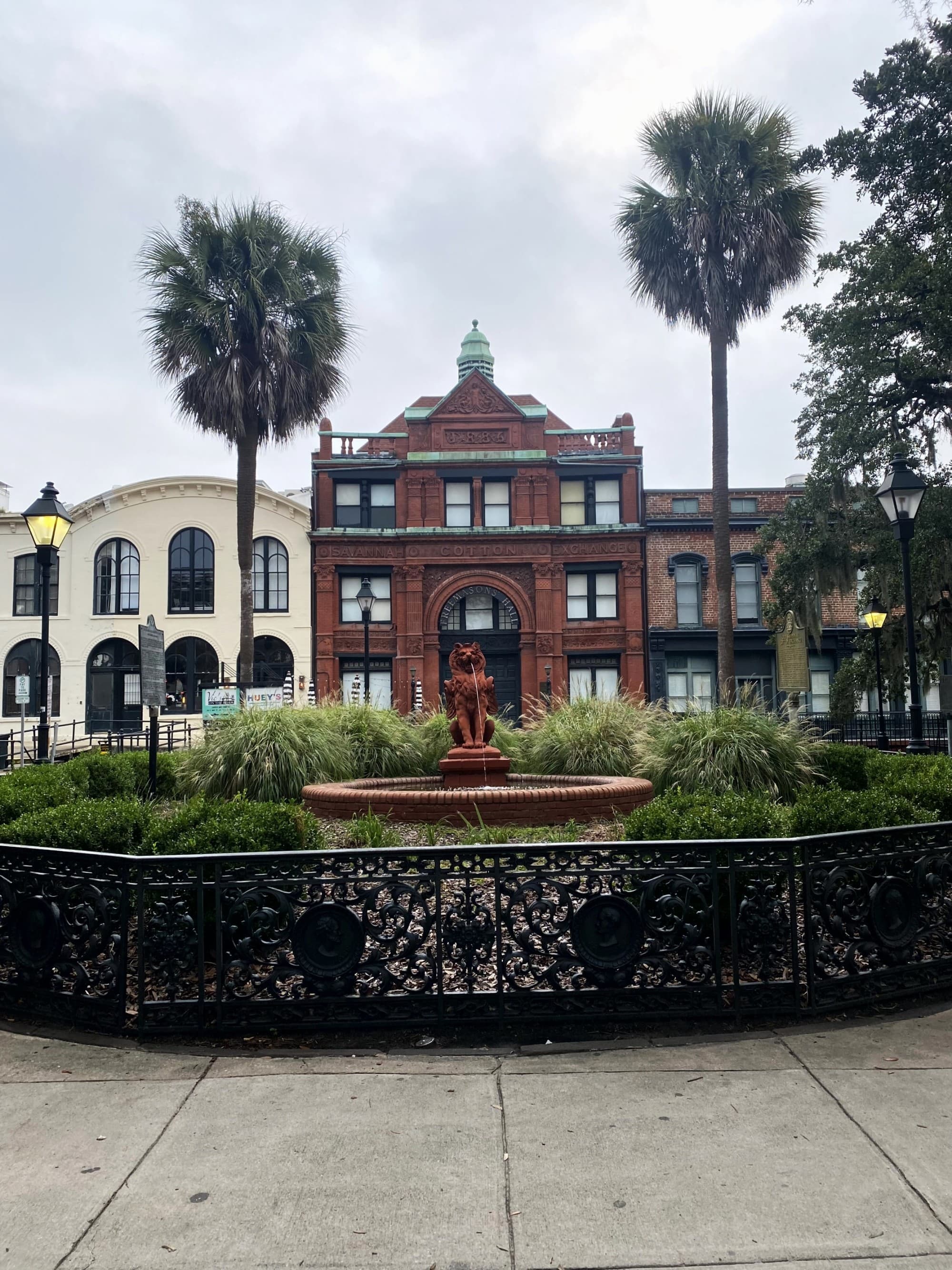 A fountain with a statue set before a red brick building with green roofing, embraced by palm trees and an iron fence, under a cloudy sky.