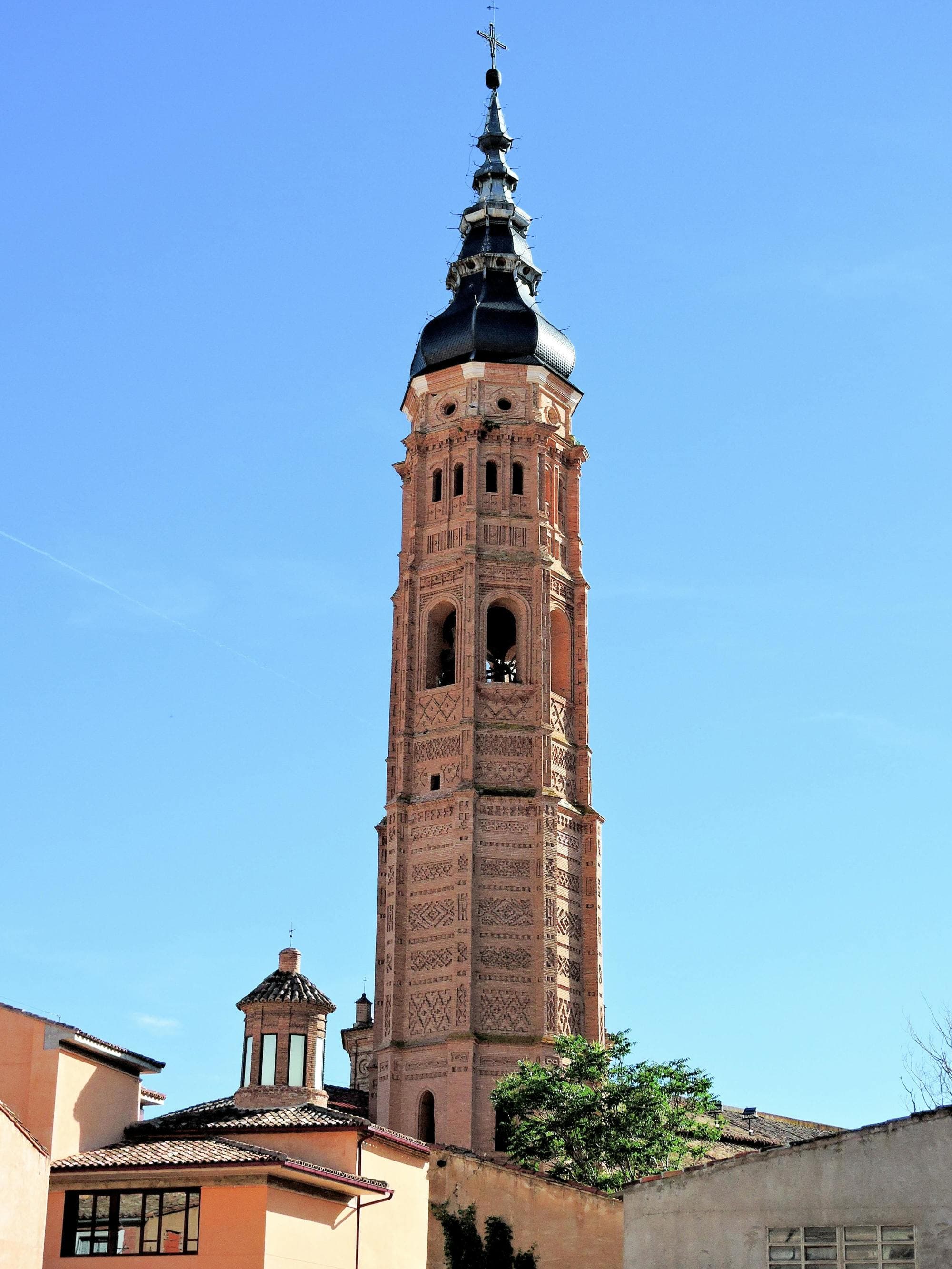 A tall ornate tower with a spire, featuring intricate brickwork patterns, set against a clear blue sky.